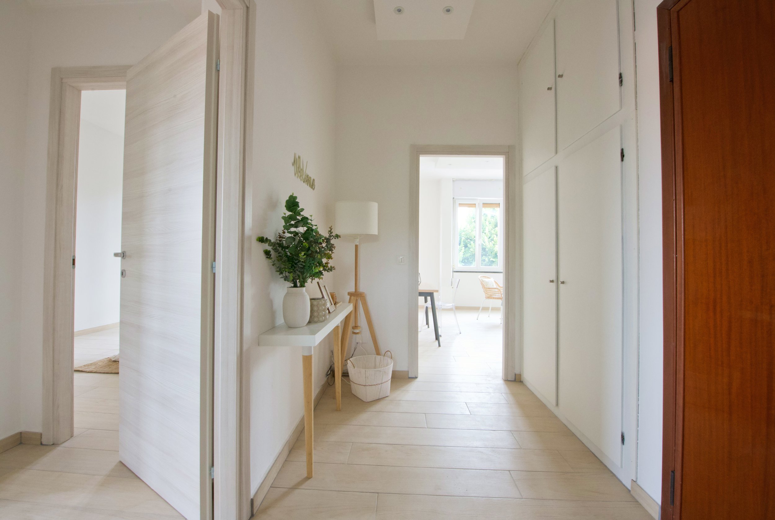 Interno di una casa moderna con corridoio luminoso, pavimento in legno chiaro, porta aperta, consolle con vaso di fiori, lampada da terra e accesso a una stanza con finestre grandi.
