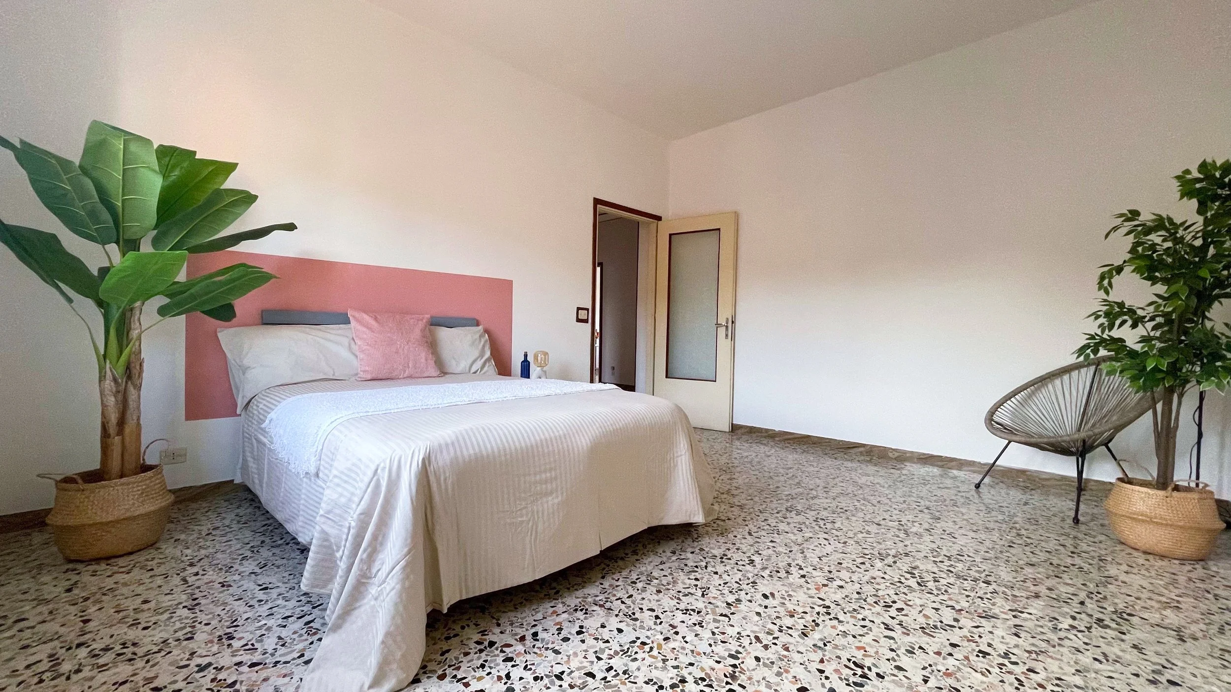 Camera da letto moderna con letto matrimoniale, parete con testiera rosa, due piante in vaso, pavimento a piastrelle e sedia a cestino nell'angolo.