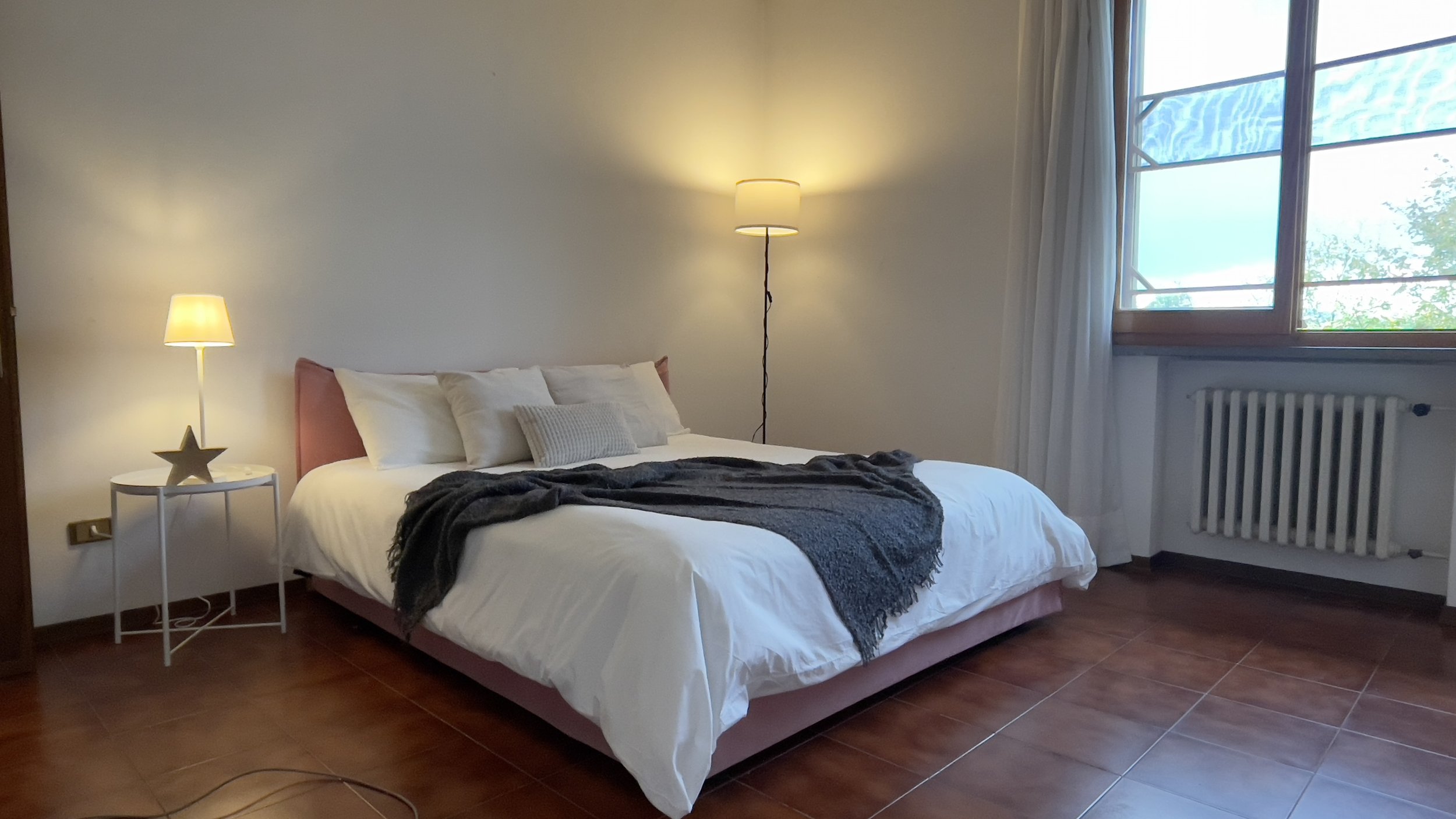 Camera da letto con letto matrimoniale, cuscini bianchi, coperta grigia, lampada da terra accesa e finestra con tende bianche. Tavolino con lampada e decorazione a forma di stella.