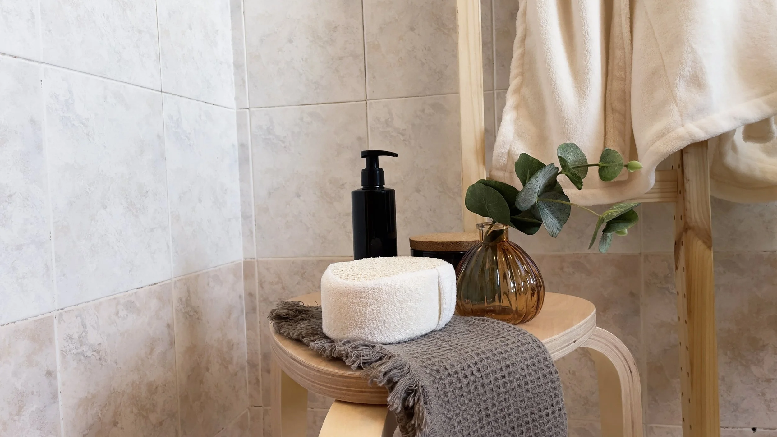 Bagno con sgabello di legno, asciugamano grigio, dispenser di sapone nero, spugna bianca e vaso con foglie verdi.