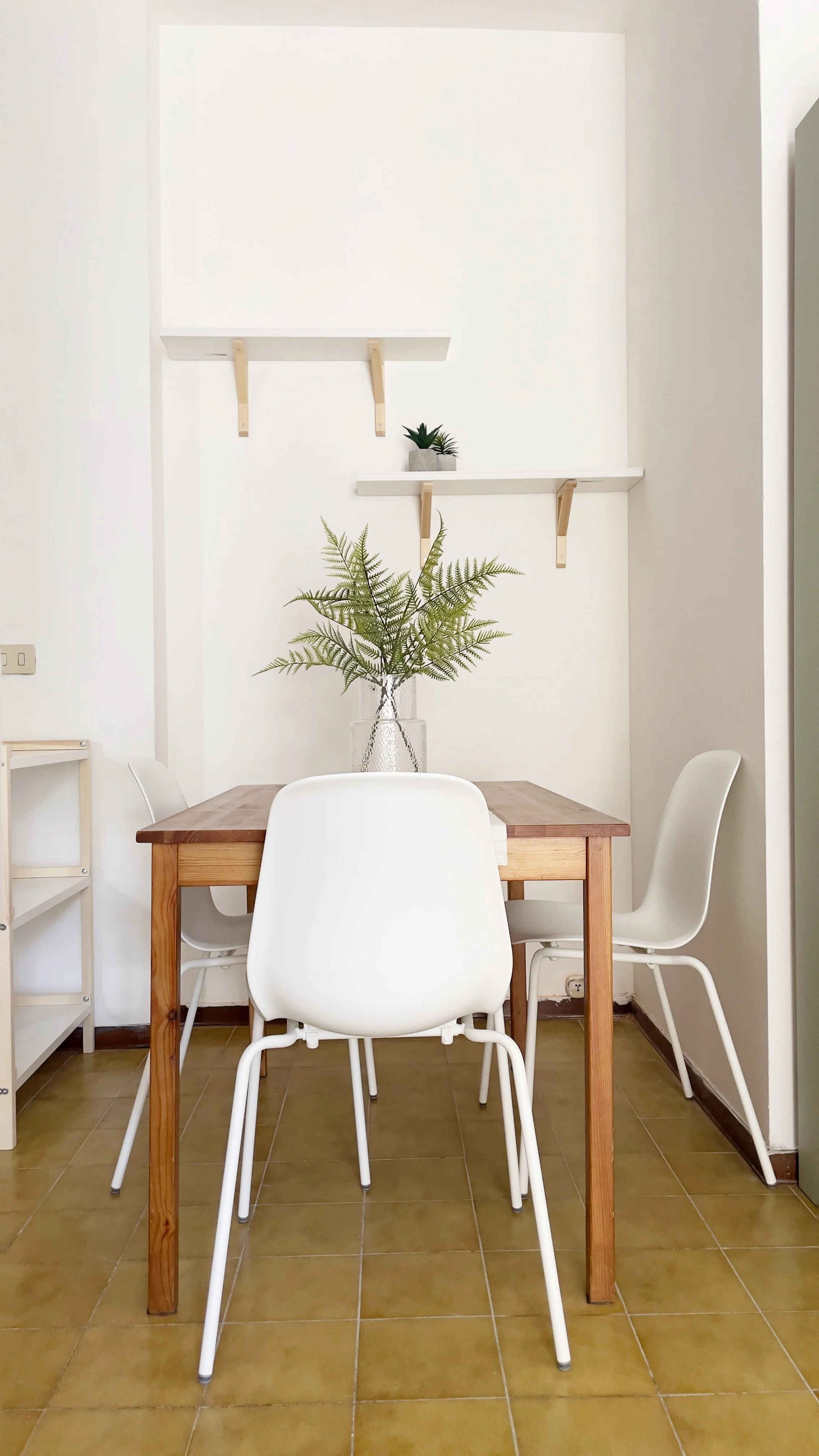Sala da pranzo luminosa con tavolo in legno, quattro sedie bianche e pianta decorativa su un vaso di vetro. Scaffali a parete con piccola pianta in vaso.