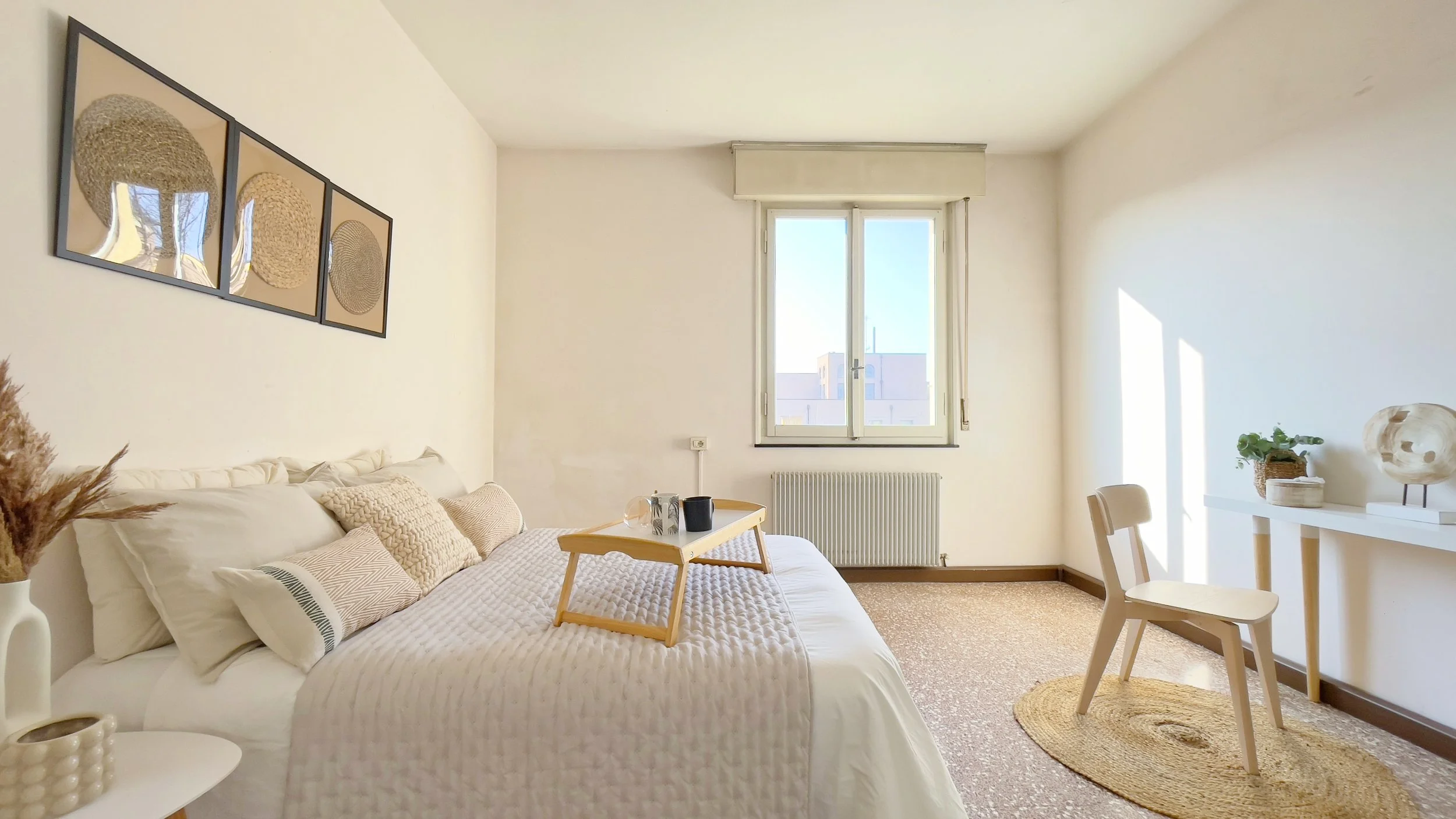 Camera da letto minimalista con letto bianco, cuscini decorativi, vassoio in legno, finestra, sedia e scrivania con piante, decorazioni naturali alle pareti.