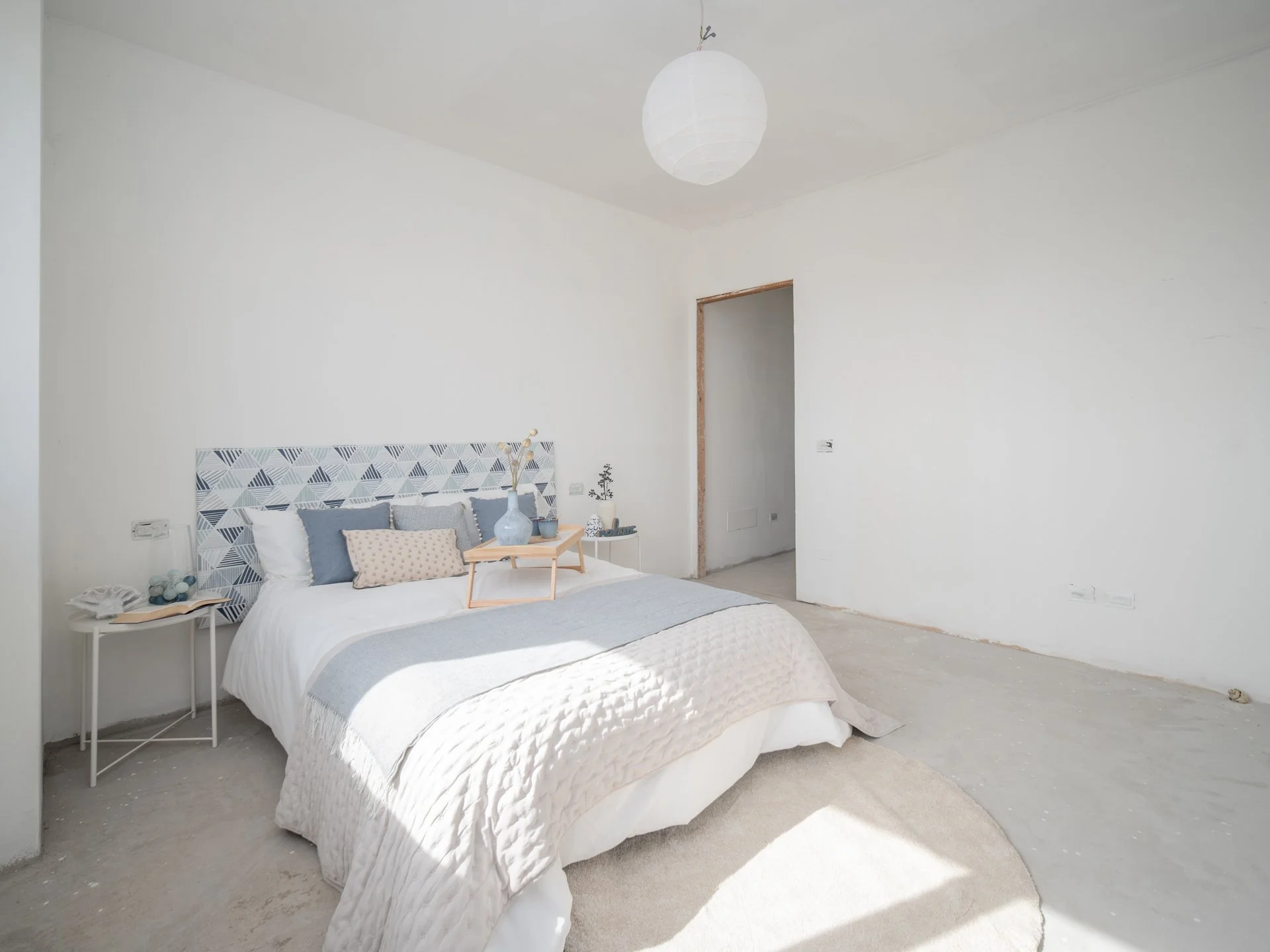 Camera da letto moderna e luminosa con pareti bianche, letto matrimoniale con copriletto bianco e cuscini decorativi, testiera geometrica in diverse sfumature di blu. Tavolino con oggetti decorativi accanto al letto e lampada a sospensione tonda bian