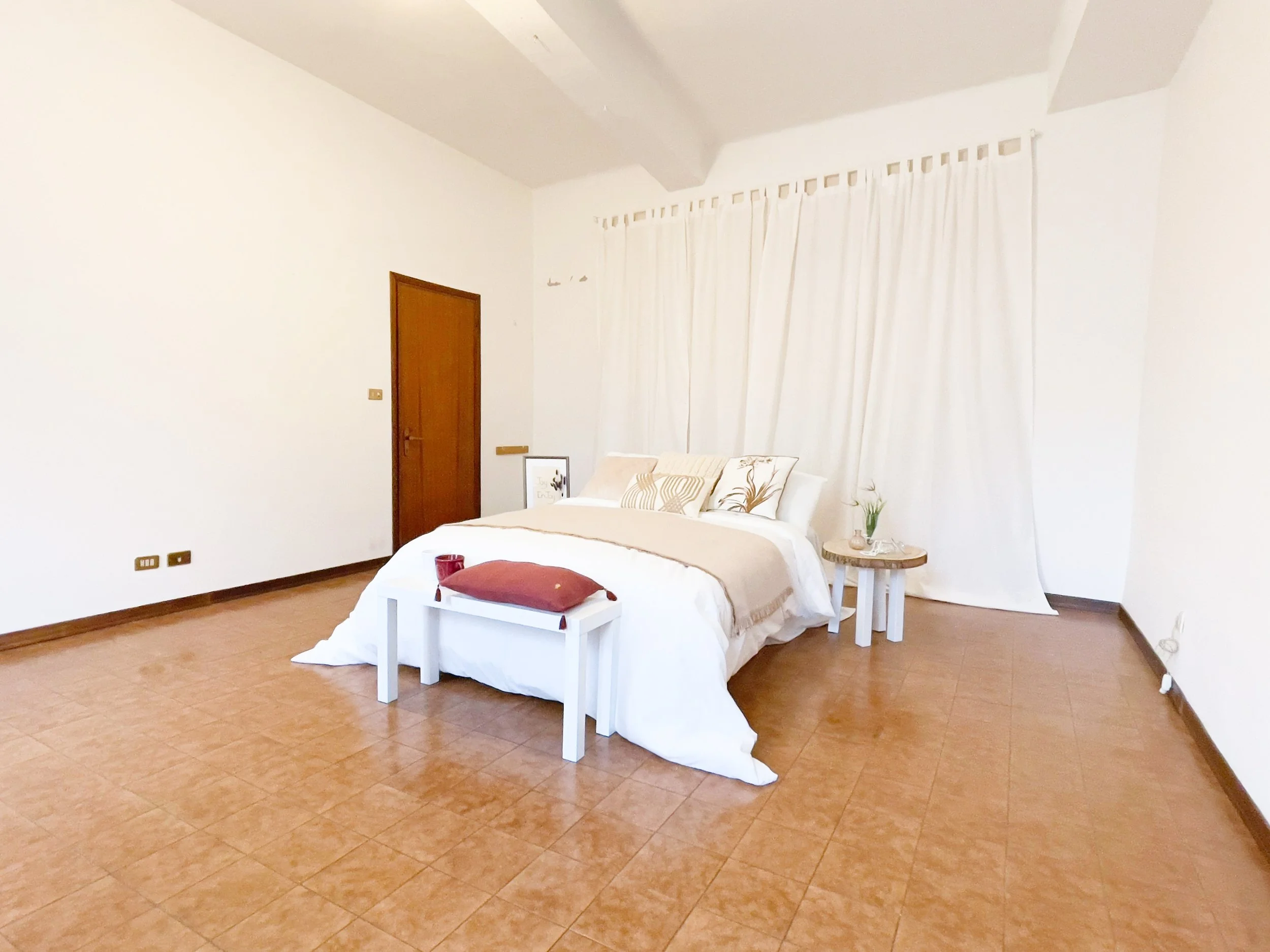 Camera da letto minimalista con letto matrimoniale, cuscini decorativi, tavolino con pianta e bicchieri, pavimento in piastrelle, porta in legno e tende bianche.