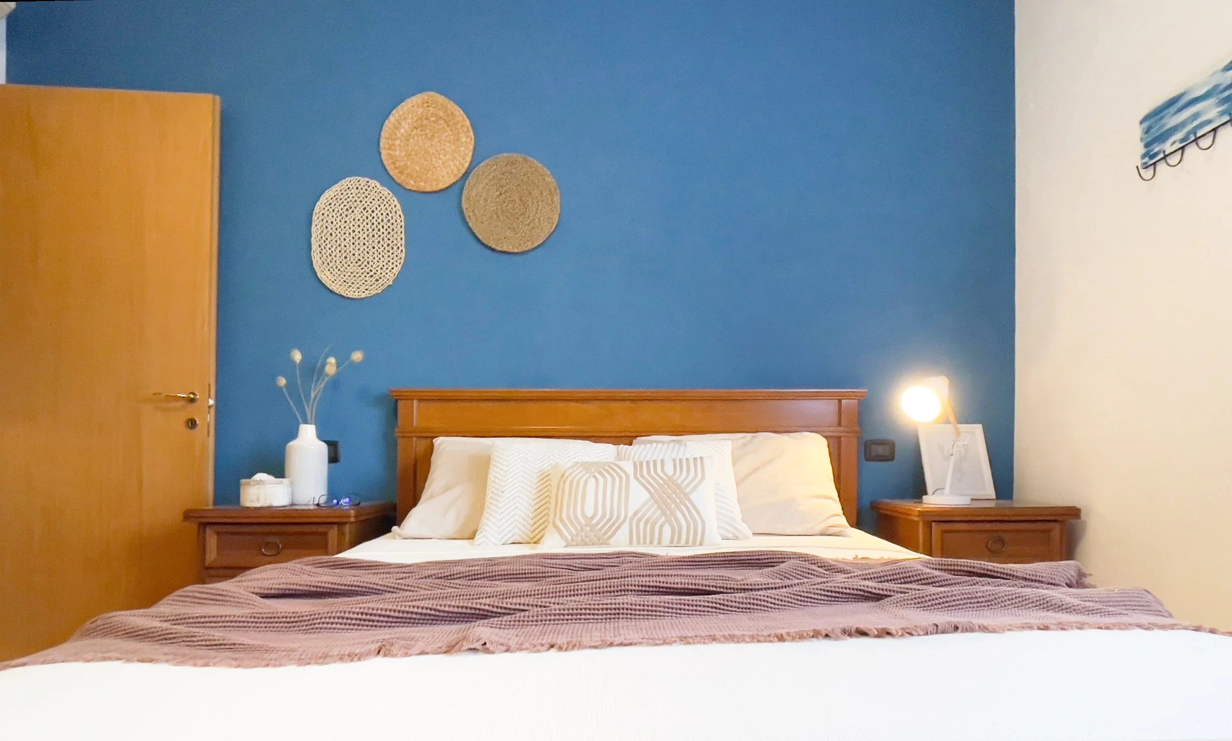 Camera da letto con parete blu, letto matrimoniale con cuscini bianchi e marroni, coperta marrone, comodini in legno con lampade e decorazioni a parete in paglia. Porta di legno aperta sulla sinistra.