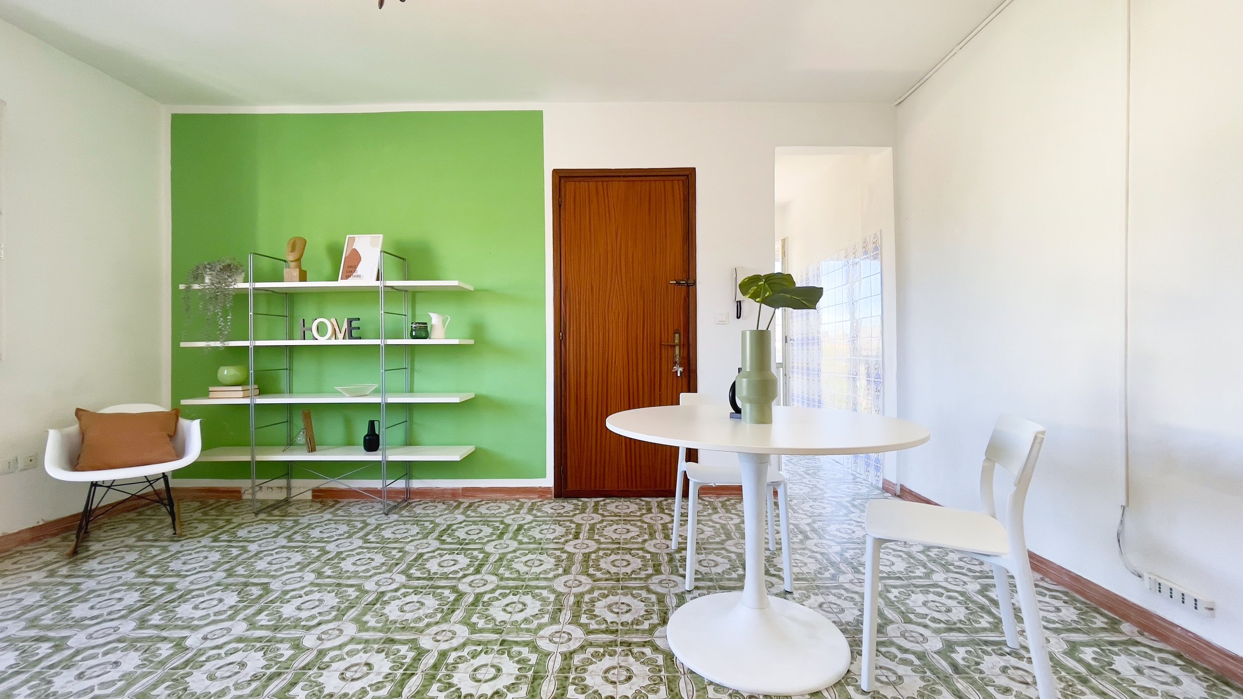 Soggiorno moderno con parete verde, porta di legno, scaffale con decorazioni, poltrona bianca con cuscino marrone, tavolo rotondo bianco con vaso e foglie, sedia bianca, pavimento con piastrelle decorato.