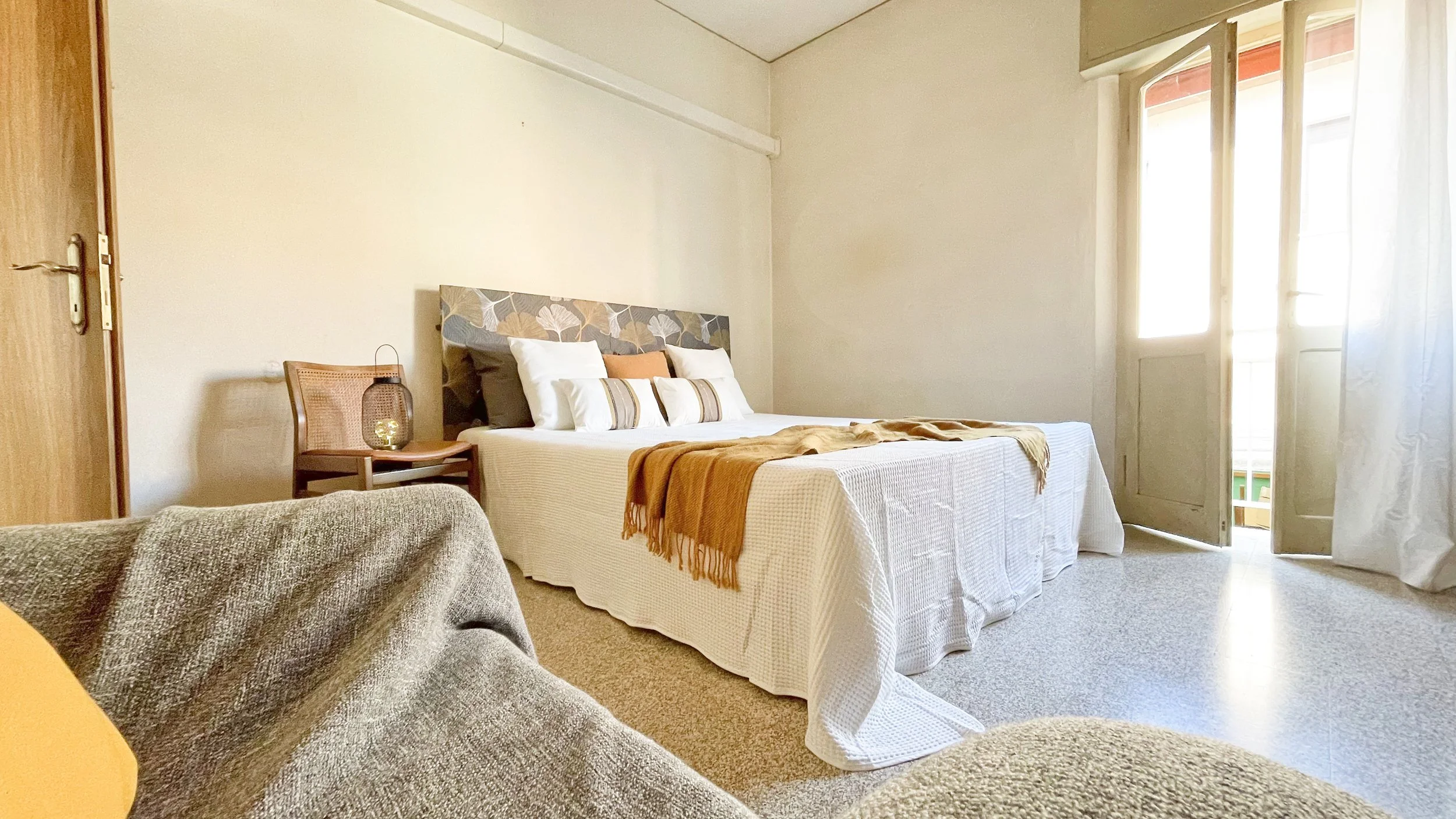 Camera da letto luminosa con letto matrimoniale, coperta beige, cuscini bianchi, testiera decorata, sedia con lampada e finestra con tenda bianca.