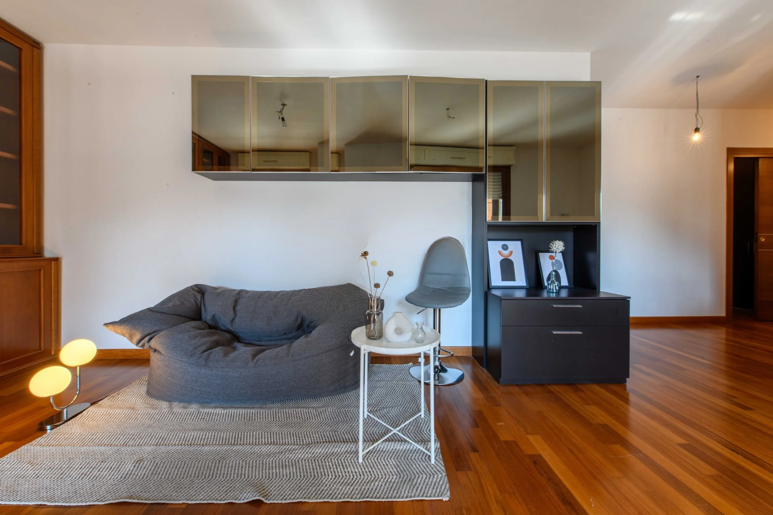 Soggiorno moderno con divano grigio, lampada da terra, tavolino bianco con vasi, pavimento in legno, mobile con specchi e decorazioni, sedia grigia e quadri astratti.