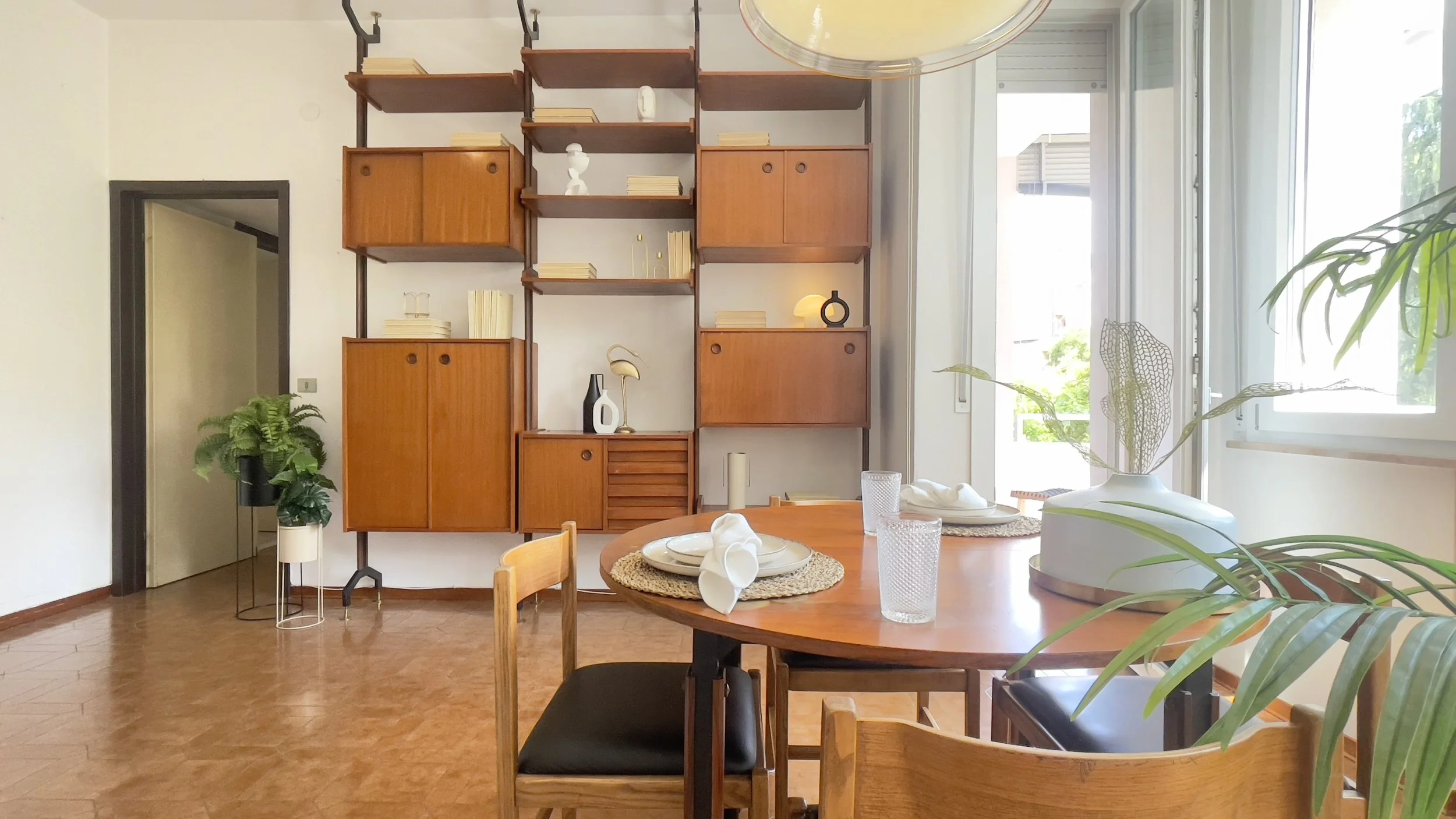 Sala da pranzo retrò con tavolo rotondo in legno apparecchiato, sedie abbinate, scaffali modulari in legno sulle pareti e piante decorative.
