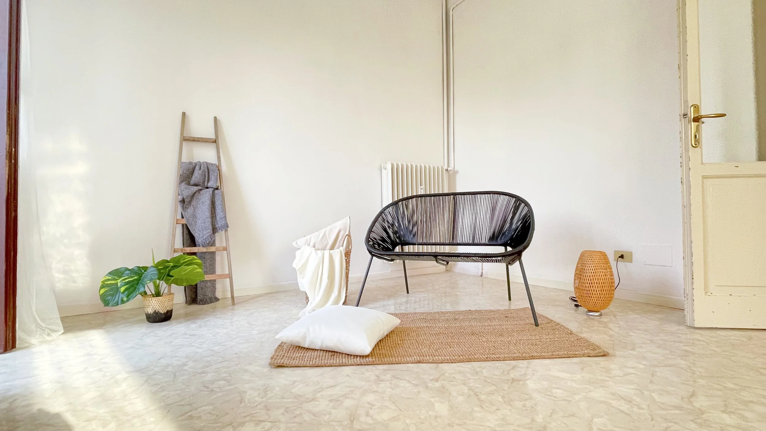 Soggiorno minimalista con sedia nera, scala in legno con coperta grigia, pianta in vaso, tappeto marrone, cuscino bianco e luce naturale.