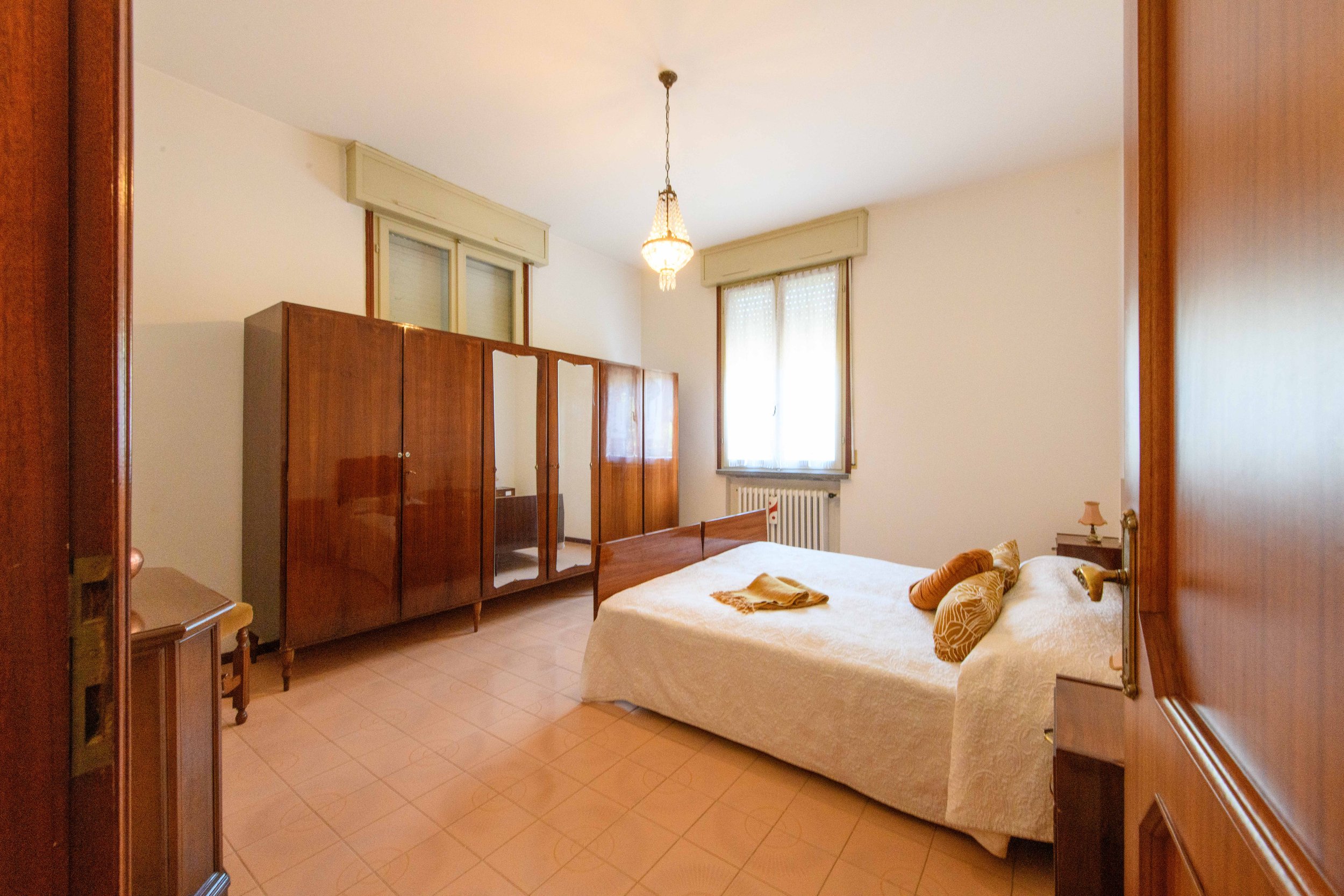 Camera da letto con letto matrimoniale, copriletto bianco e cuscini decorativi marroni, grande armadio con ante in legno e specchi, lampadario appeso al soffitto, e finestra con tende bianche; pavimento piastrellato.