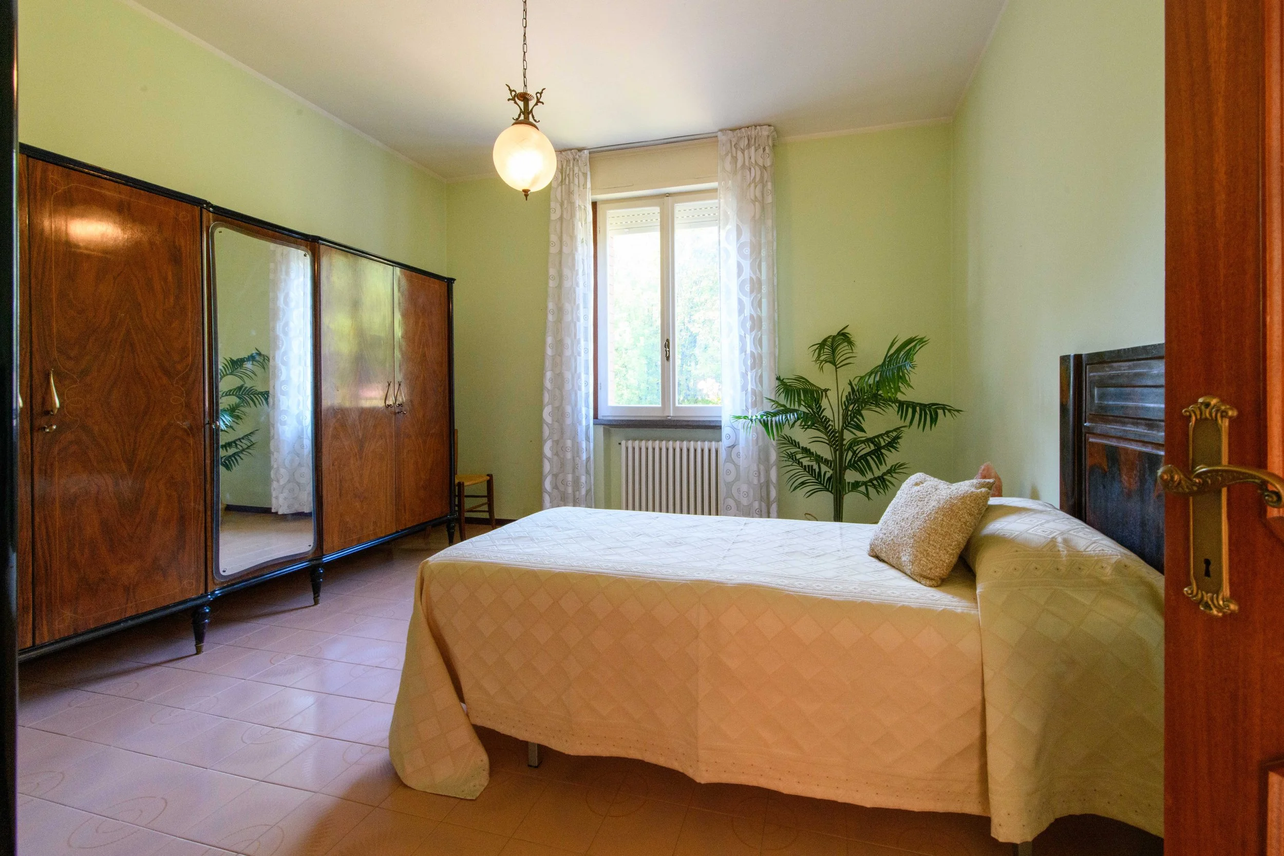 Camera da letto con letto singolo, copriletto beige, armadio in legno, finestra con tende bianche a motivi, pianta decorativa e lampadario sospeso.