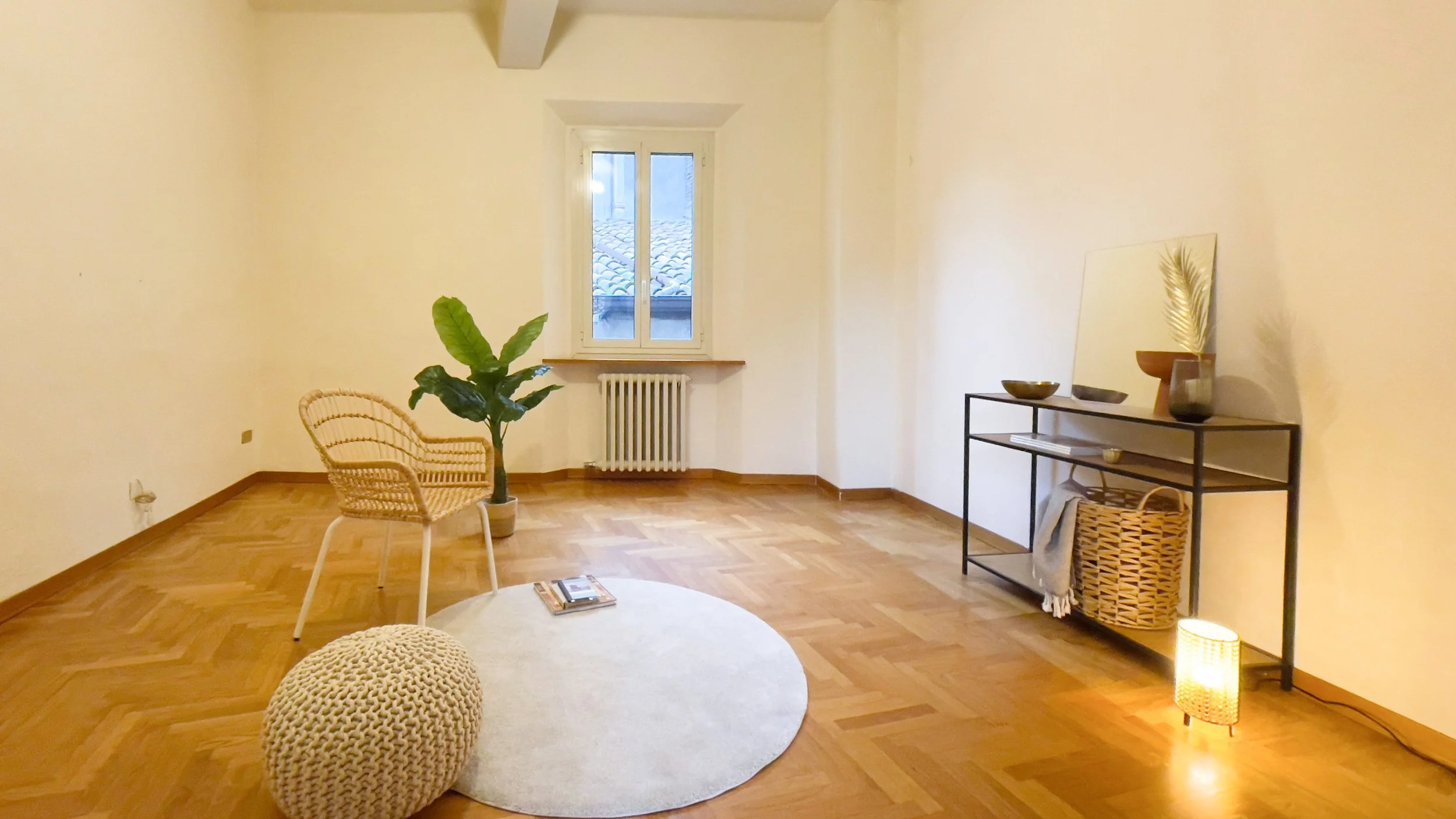 Soggiorno minimalista con sedia in vimini, pianta verde, tappeto circolare, pouf lavorato a maglia, finestra, consolle con decorazioni e lampada accesa su pavimento in legno.