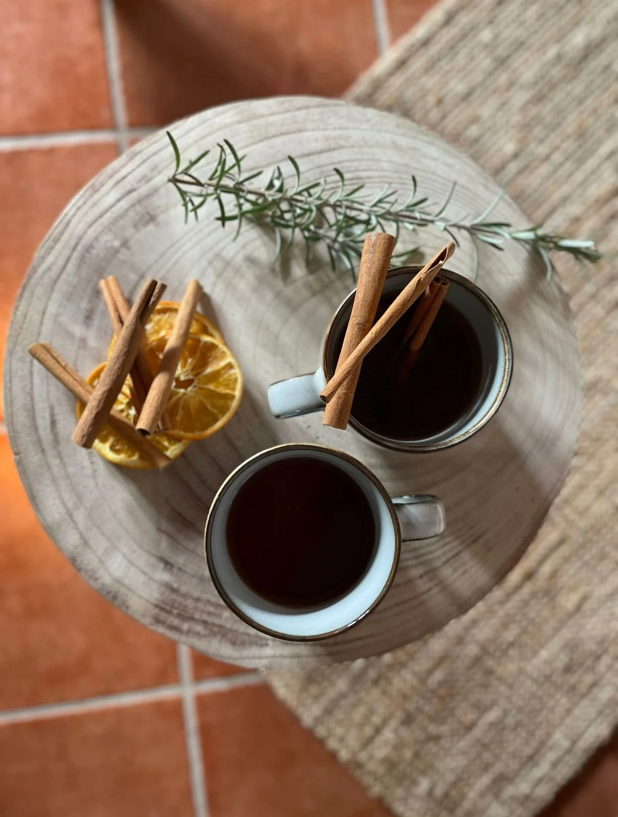 Due tazze di tè con bastoncini di cannella su un tavolino rotondo, decorato con rametto di rosmarino e fette d'arancia essiccate, su pavimento di piastrelle.