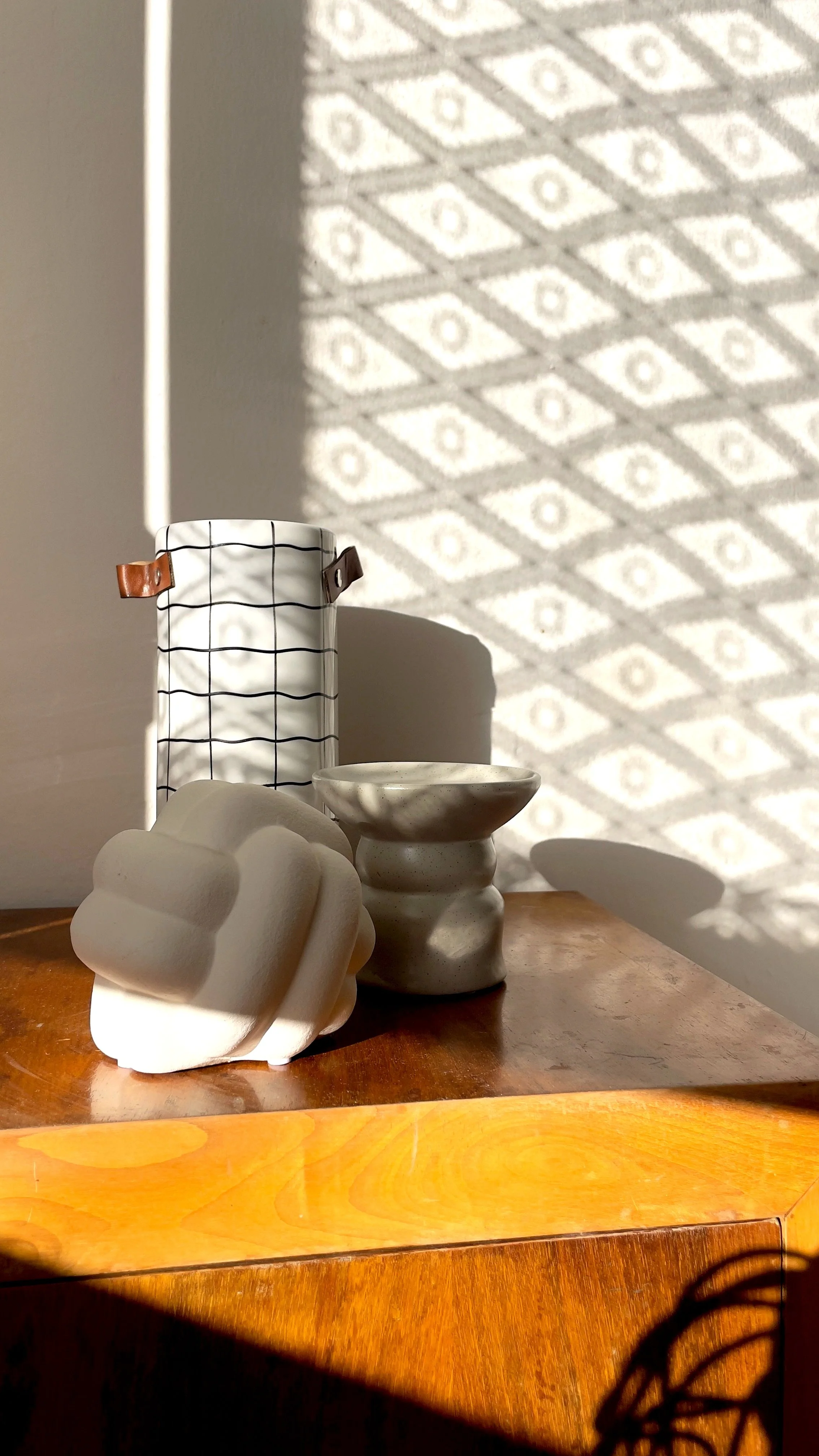 Oggetti decorativi su un tavolo di legno, incluso un vaso a scacchi con dettagli in pelle, una decorazione geometrica e un piccolo contenitore. Sul muro si riflette un pattern di ombre esagonali.