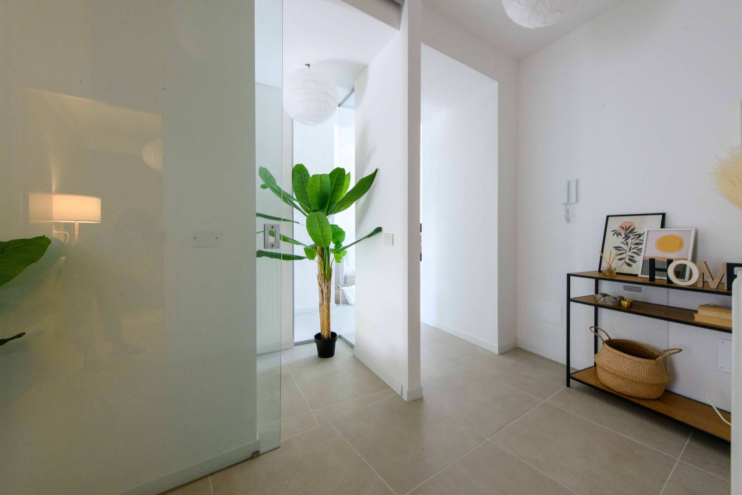 Interno di una casa moderna con pareti bianche, pavimento in piastrelle chiare, una pianta tropicale in un vaso nero e una mensola con decorazioni e un cesto di vimini.