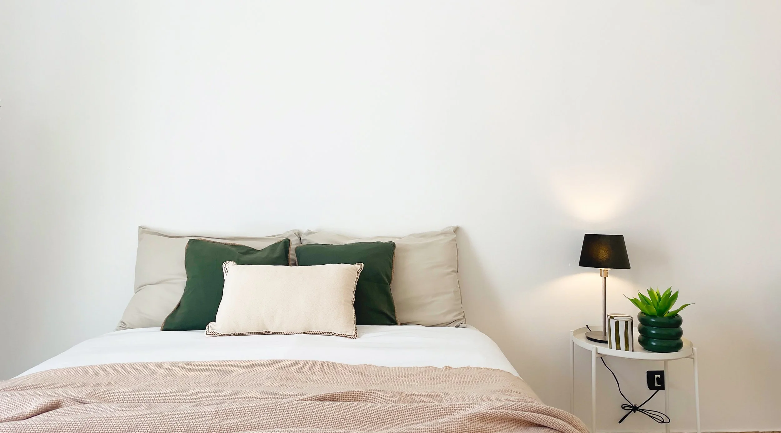 Camera da letto minimalista con letto, cuscini verdi e beige, lampada da tavolo nera, e pianta su comodino bianco.