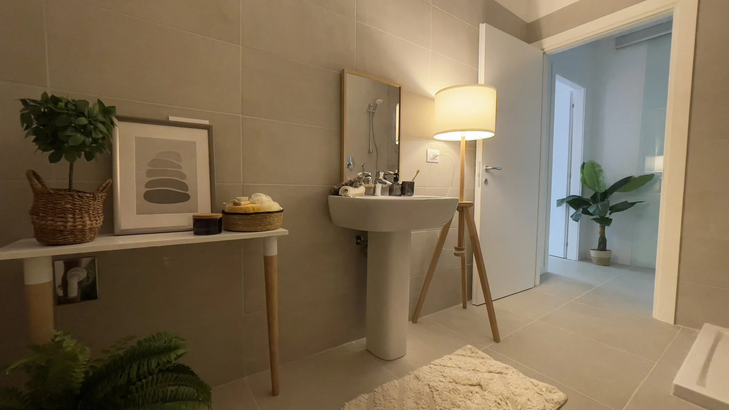Interno di un bagno moderno con lavabo, specchio, lampada da terra, piante, e decorazioni su un tavolino. Porta aperta conduce a un'altra stanza. Design minimalista e accogliente.