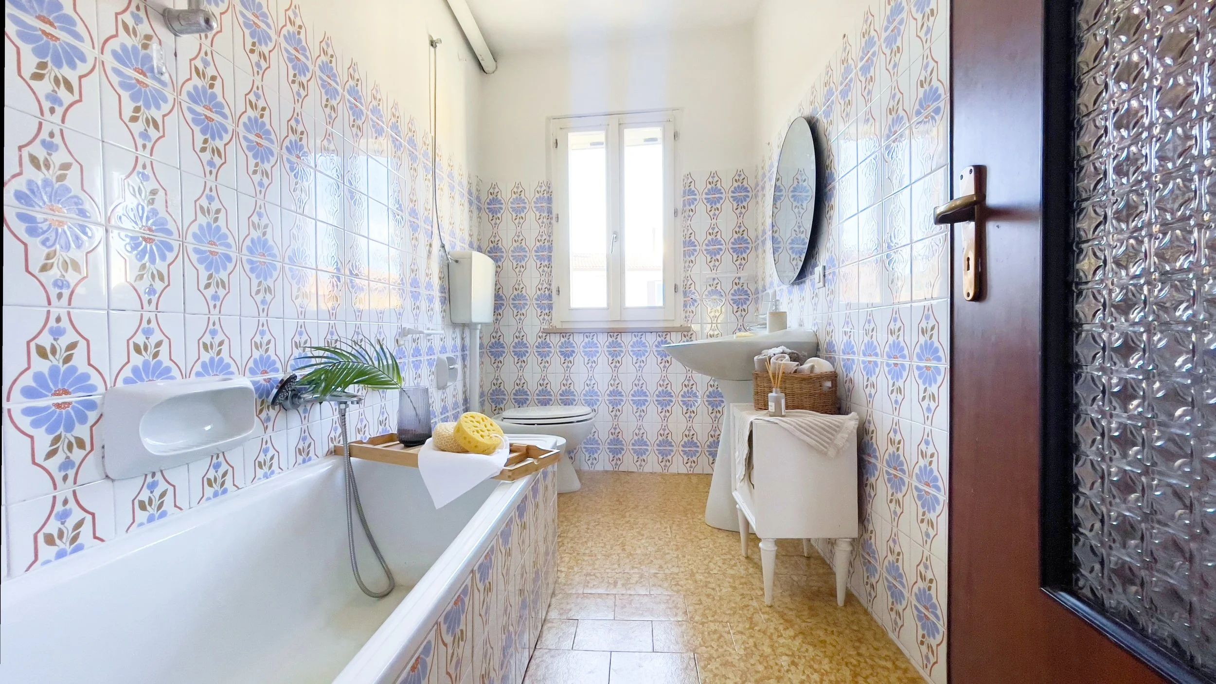 Bagno con piastrelle decorative blu e bianche, vasca da bagno, lavandino, finestra, e porta. Accessori in legno e cesto di prodotti da bagno sono visibili.