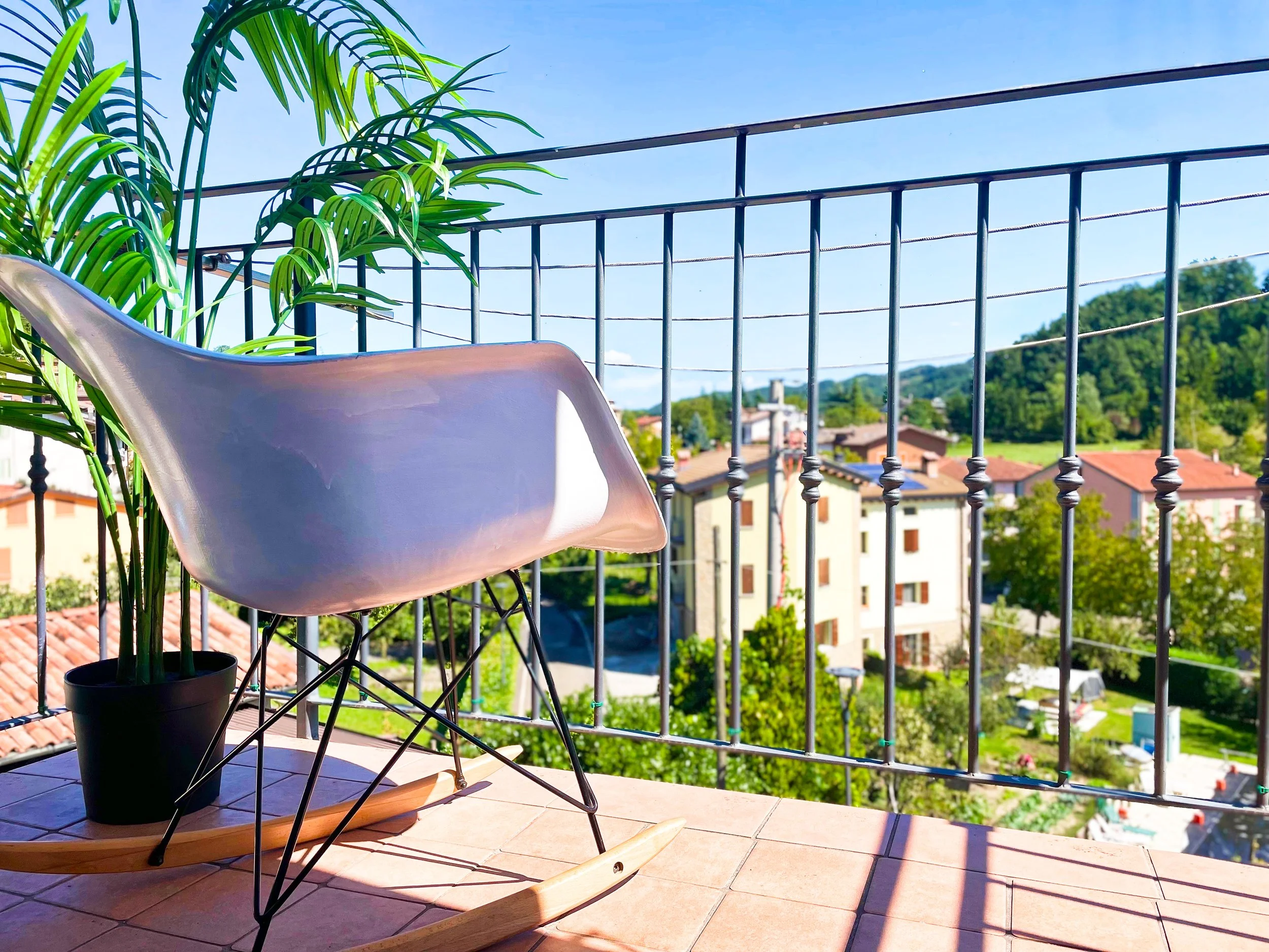 Sedia a dondolo bianca su un balcone con vista sulla città e colline verdi, pianta in vaso accanto.