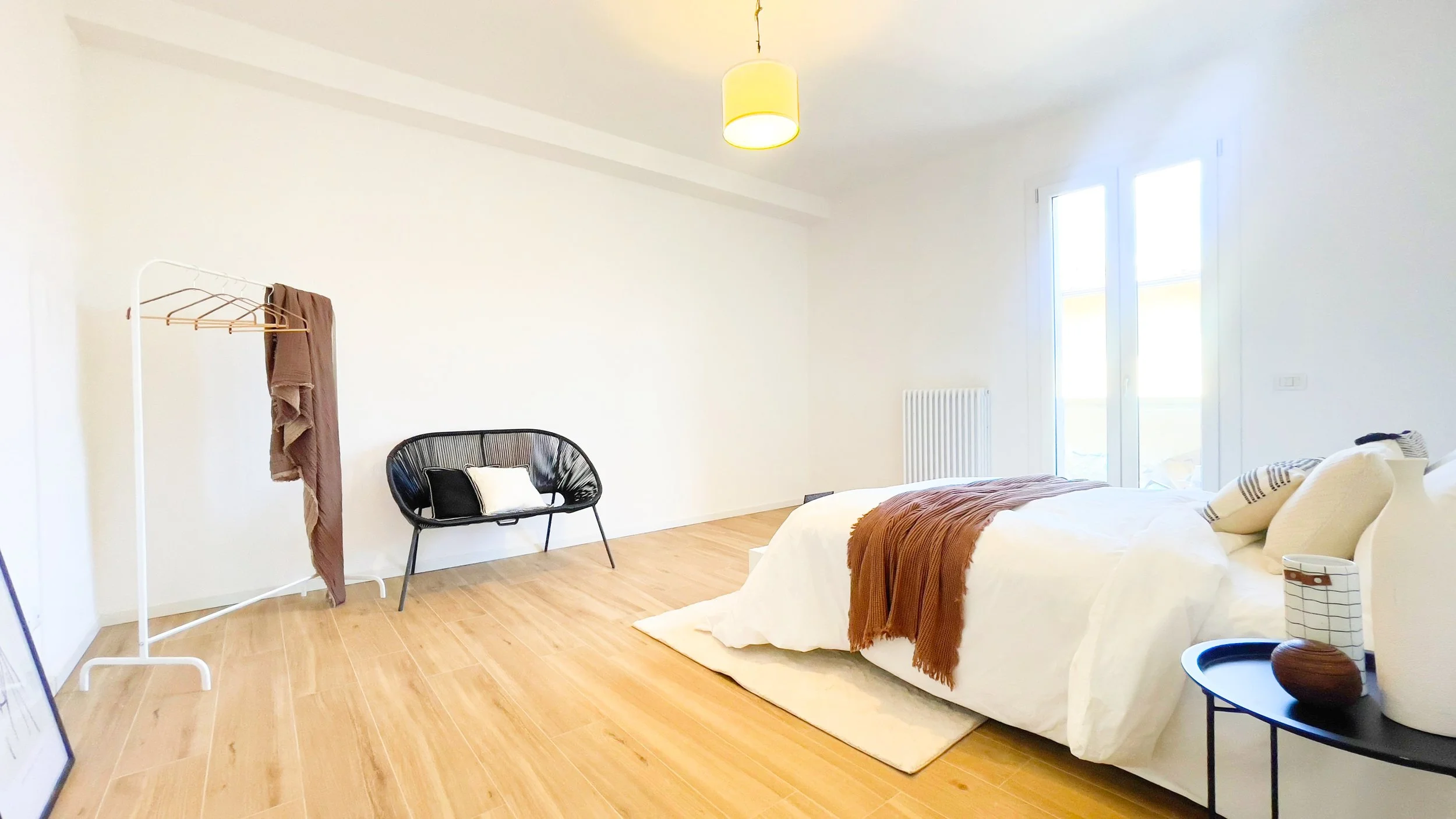 Camera da letto moderna con letto matrimoniale, coperta marrone, poltrona nera, appendiabiti bianco, pavimento in legno chiaro e finestra luminosa.