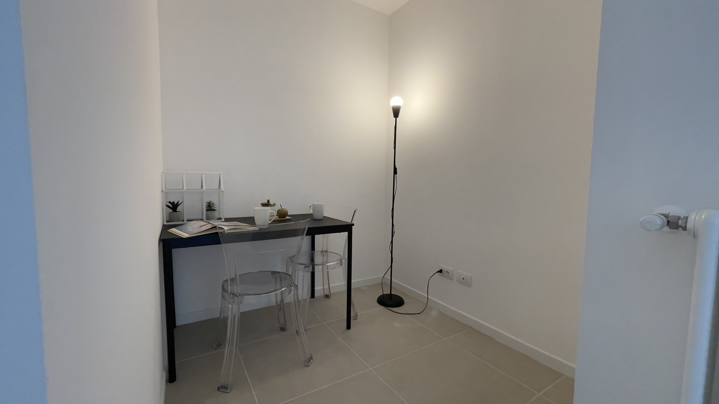 Angolo ufficio con tavolo nero, due sedie trasparenti, lampada da terra, pianta decorativa, tazza e libri su pavimento piastrellato beige.