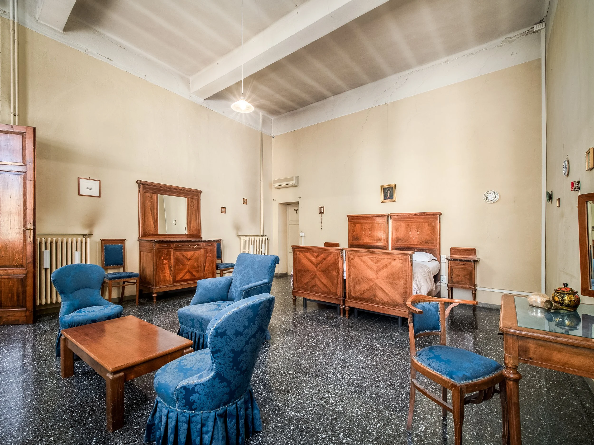 Camera con mobili in legno scuro, due letti singoli, poltrone e sedie rivestite in tessuto blu, tavolino basso e specchio. Pareti chiare e pavimento scuro.
