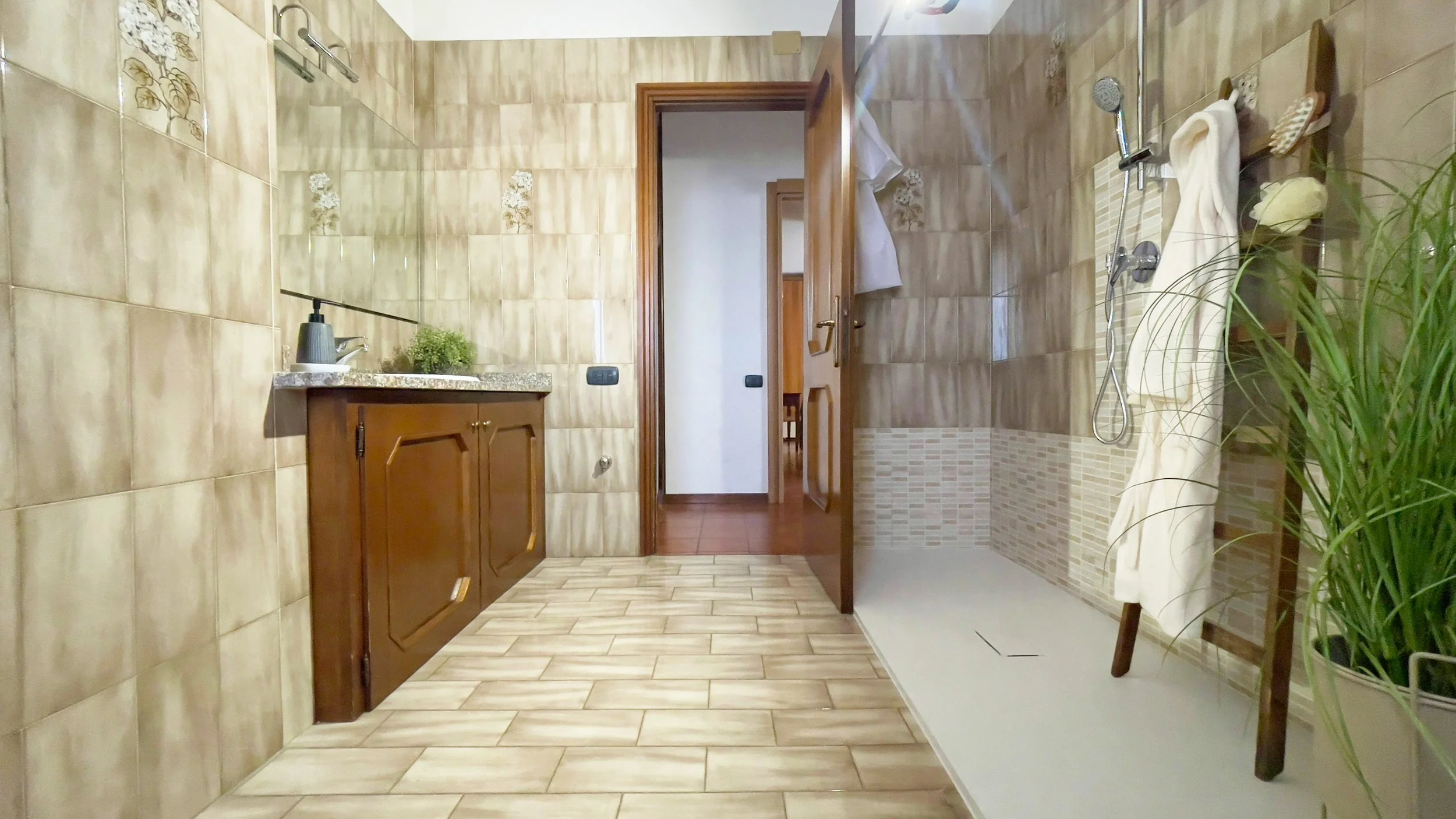 Bagno con pareti piastrellate beige, doccia con rubinetto e soffione, lavabo con armadietto in legno, specchio e pianta decorativa.