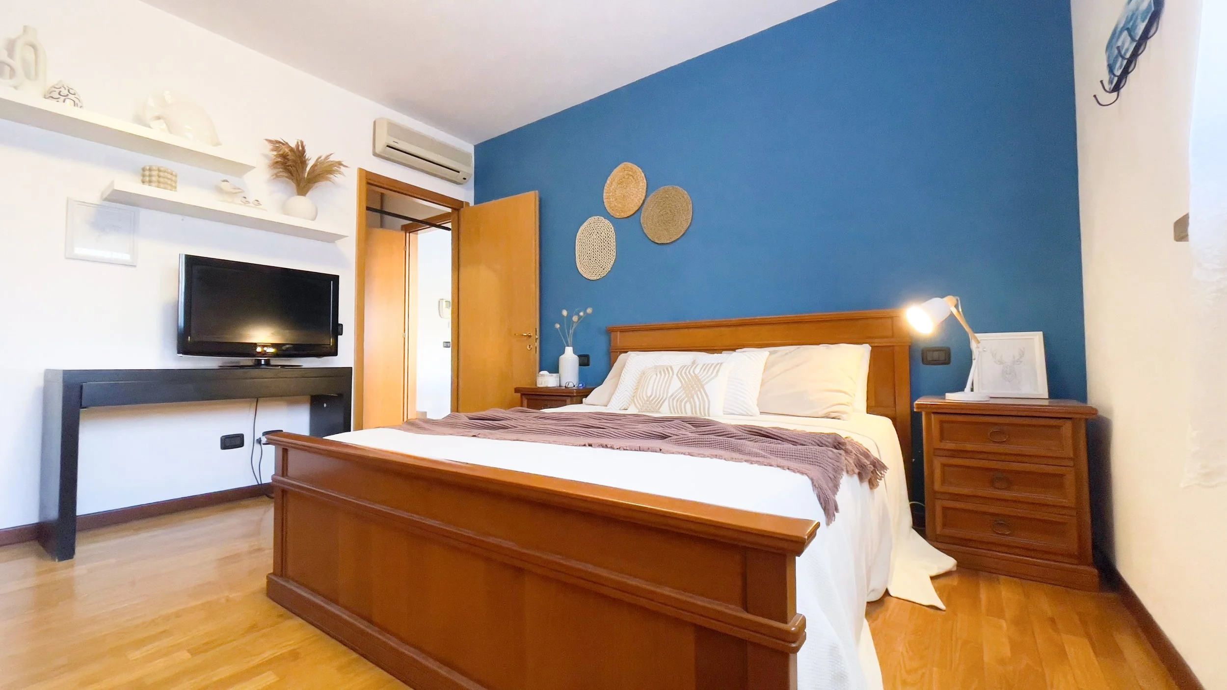 Camera da letto con parete blu, letto matrimoniale in legno, comodino con lampada, e TV su un mobile. Decorazioni a parete e scaffali con oggetti decorativi.