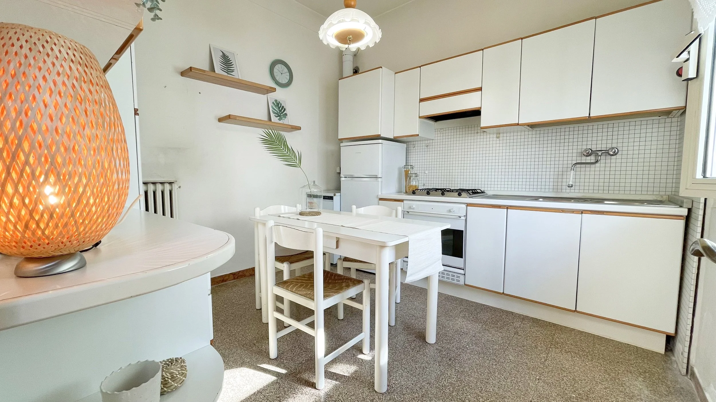 Cucina con mobili bianchi, tavolo con sedie, lampada intrecciata accesa, mensole con decorazioni, e frigorifero bianco.