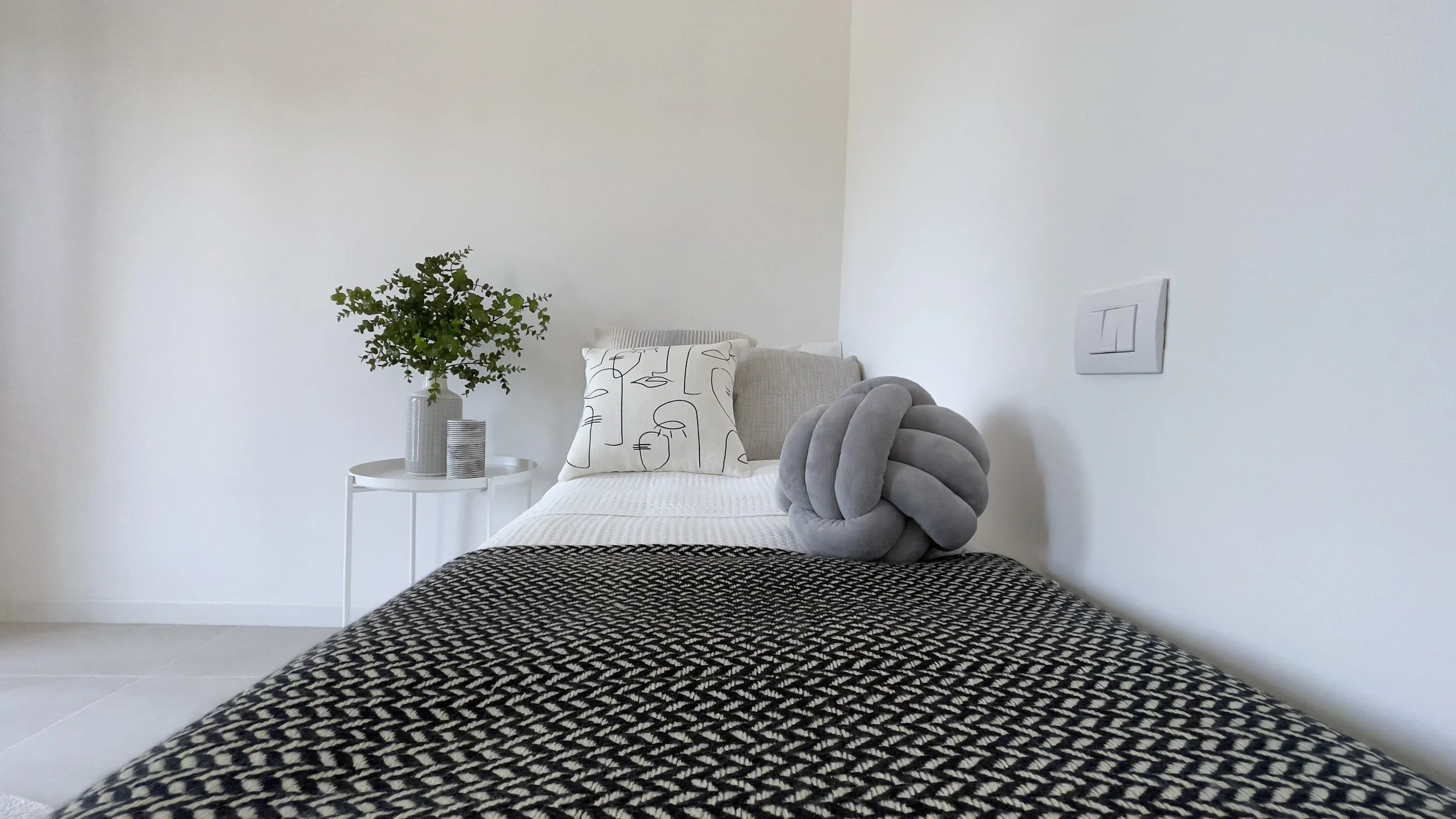 Letto singolo con cuscini, coperta decorativa e tavolino con pianta in vaso.