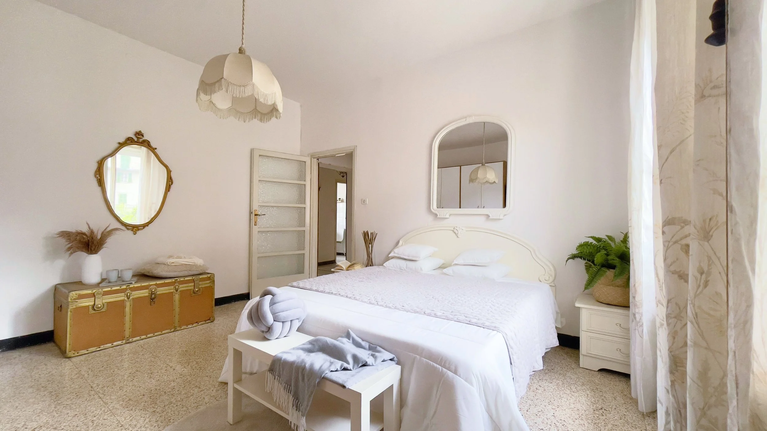 Camera da letto luminosa con letto matrimoniale, pareti bianche, specchio d'epoca, lampada a sospensione, bauli e decorazioni naturali.