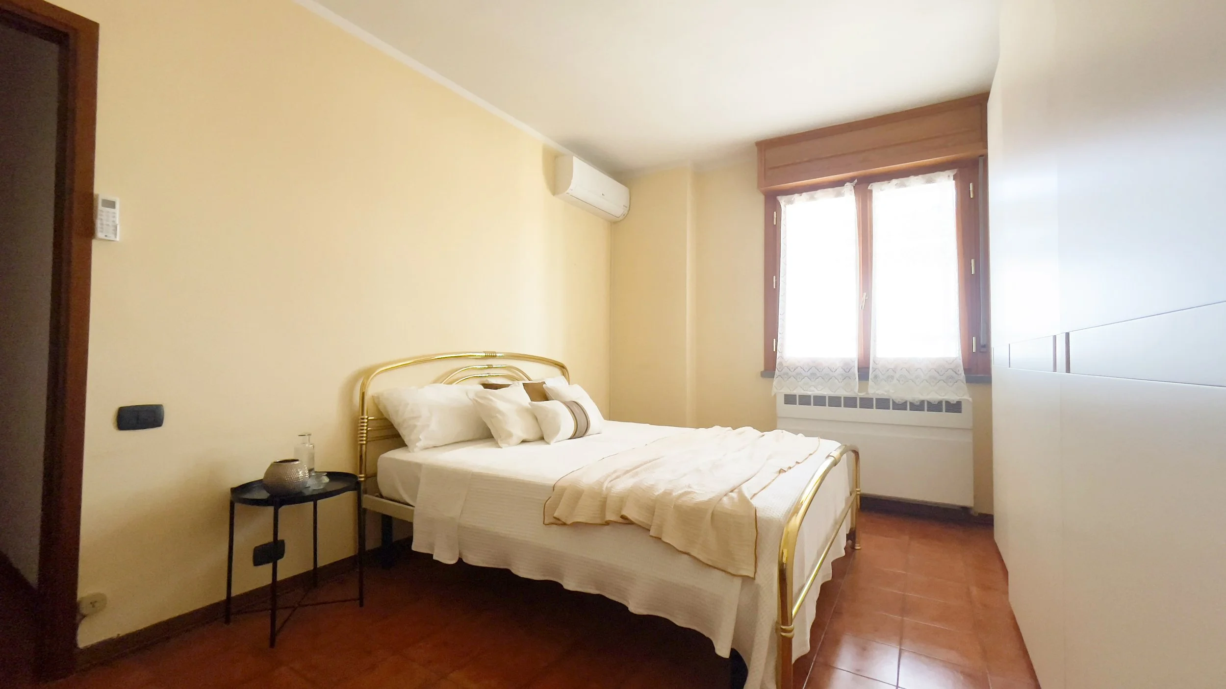 Camera da letto con letto matrimoniale, testiera in metallo dorato, comodino nero con vaso, pareti color crema, condizionatore d'aria, finestra con tende bianche e armadio bianco.