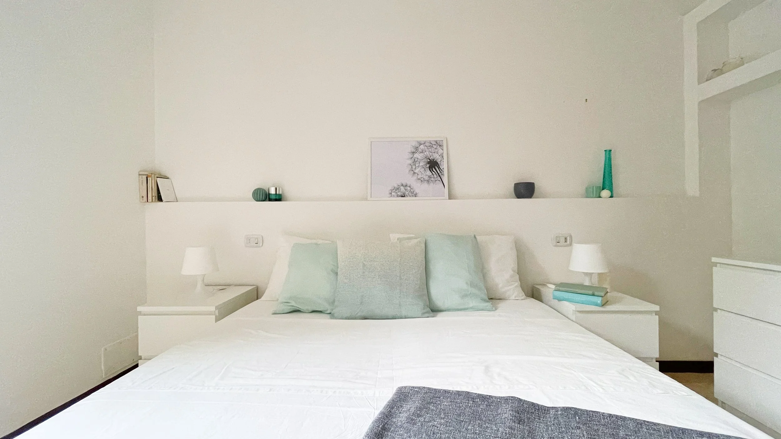 Camera da letto moderna con letto matrimoniale bianco, cuscini verde chiaro, comodini bianchi con lampade, e decorazioni minimaliste sul ripiano dietro il letto. Immagine di soffioni appesa al muro.