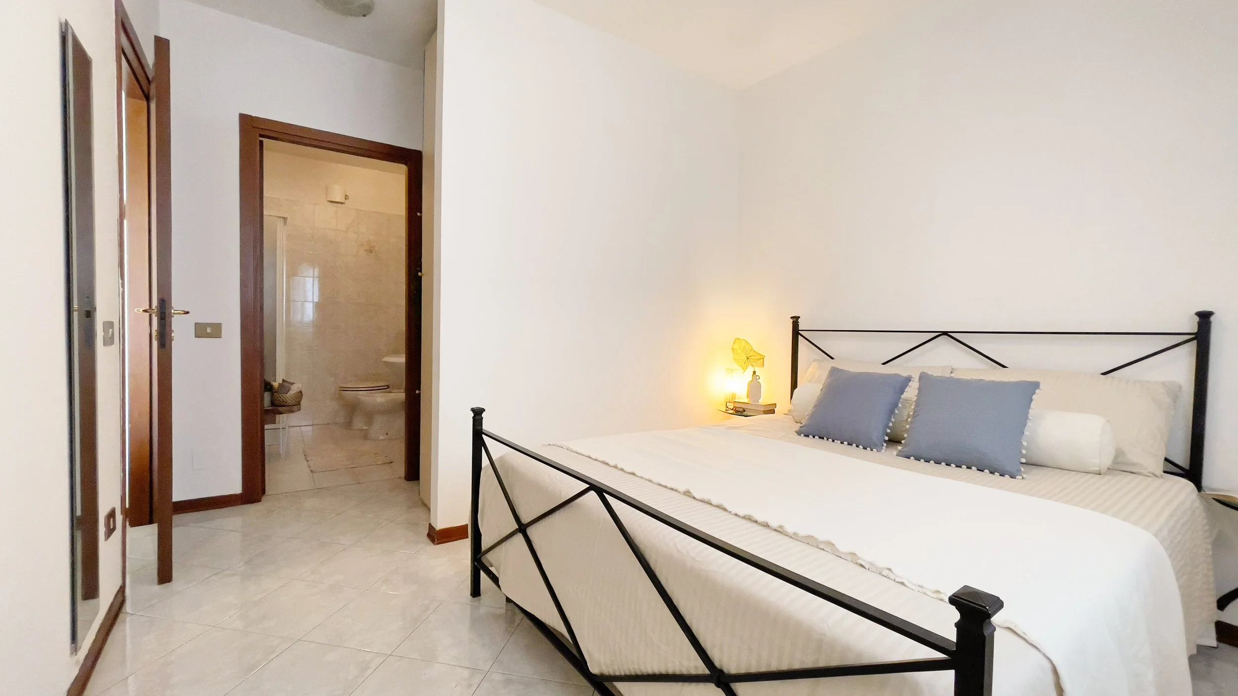 Camera da letto luminosa con letto matrimoniale, cuscini blu, comodino con lampada accesa, pavimento piastrellato chiaro, porta aperta verso un bagno con sanitari visibili.