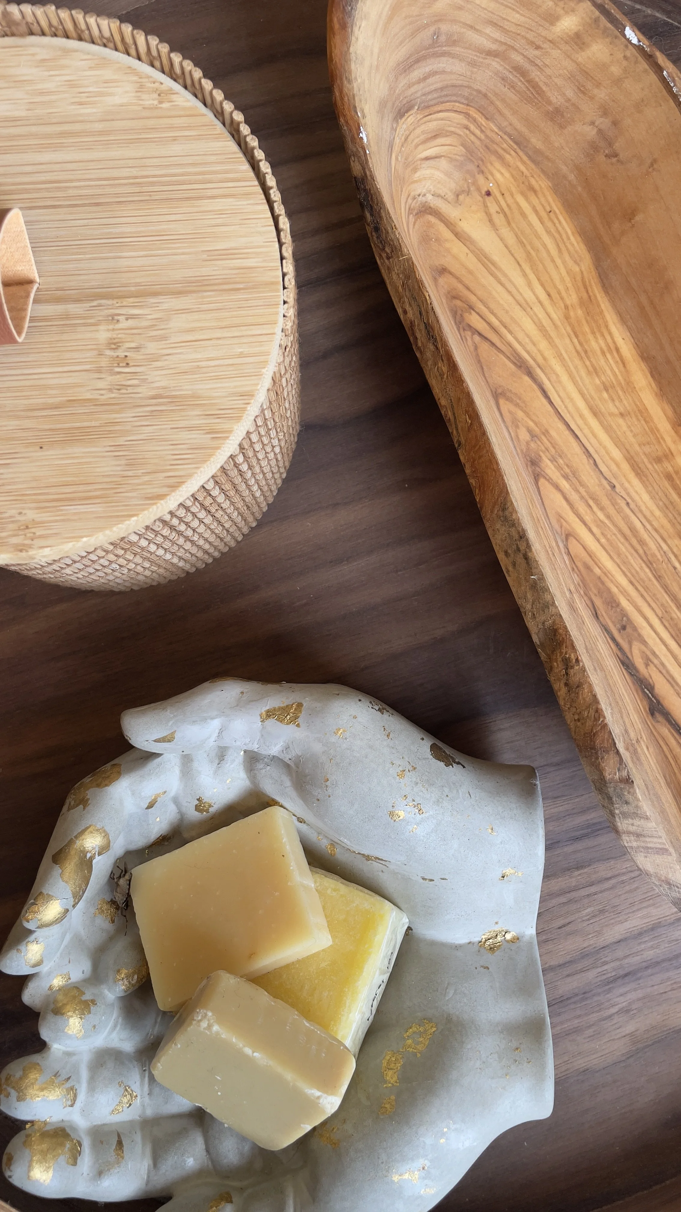 Scultura di mani che sorregge saponette, ciotola di legno e contenitore di bambù su tavolo di legno.