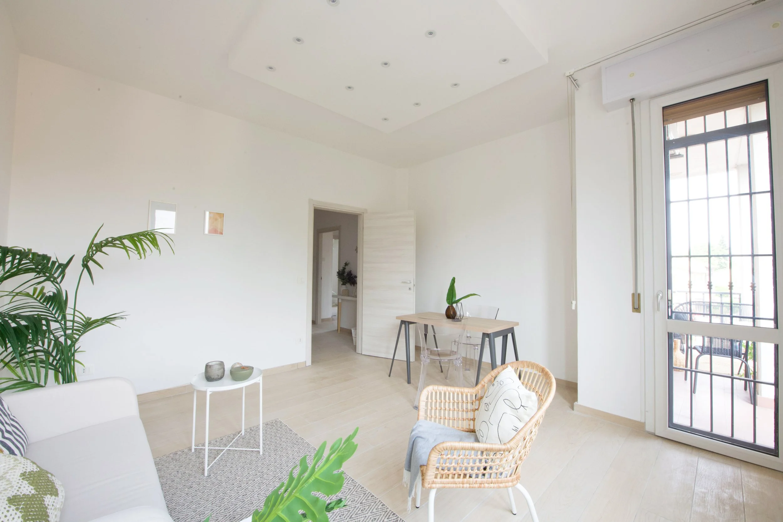 Soggiorno moderno e luminoso con divano bianco, sedia di vimini con cuscino e coperta, tavolino bianco, piante verdi e tavolo da pranzo in legno. Pareti bianche e pavimento in legno chiaro, con una grande finestra che si affaccia su un balcone. Arred