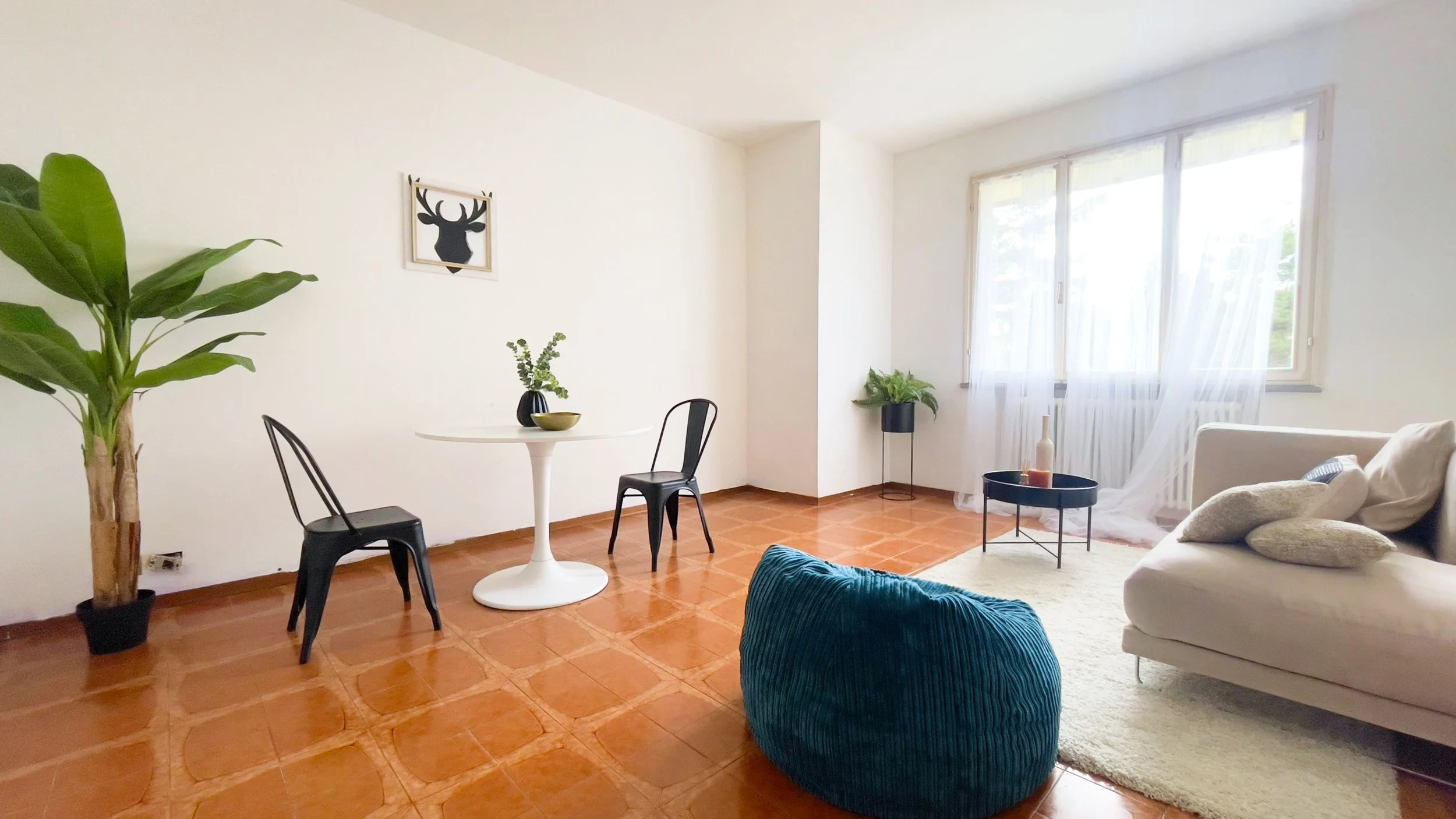 Soggiorno minimalista con pareti bianche, pavimento in piastrelle marroni, tavolo bianco con due sedie nere, pianta in vaso, divano beige con cuscini e poltrona blu. Finestra grande con tende bianche.