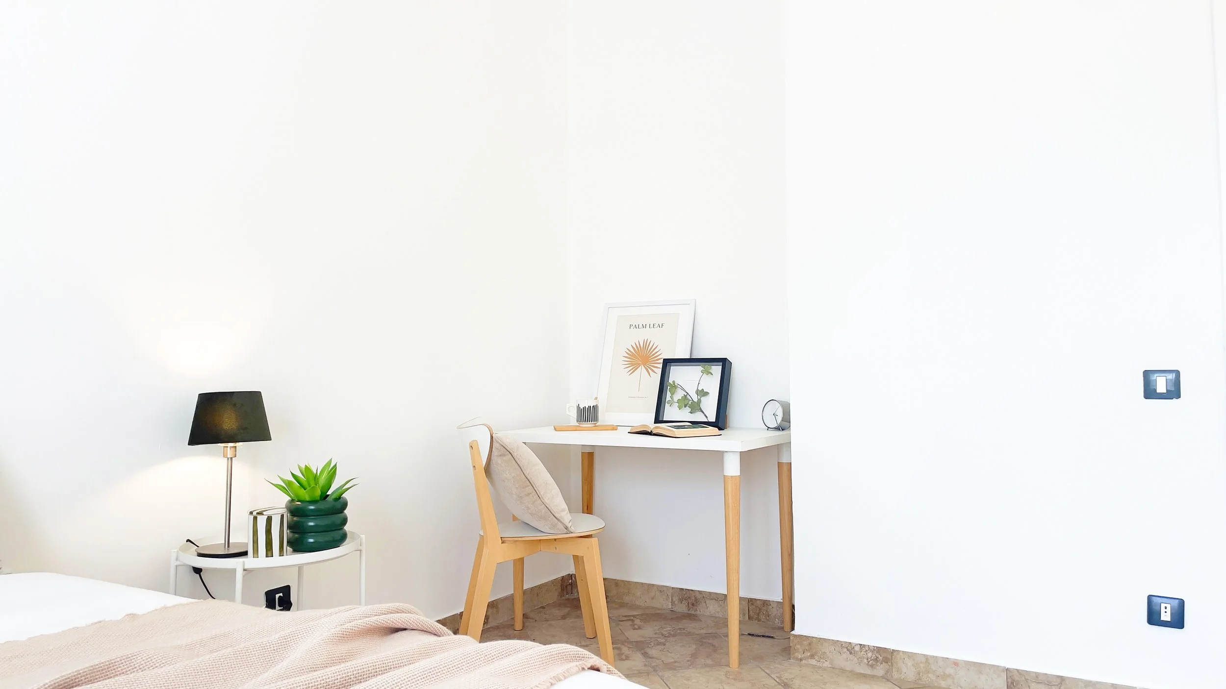Camera da letto moderna con scrivania bianca, sedia con cuscino, lampada, pianta e decorazioni sulla parete.