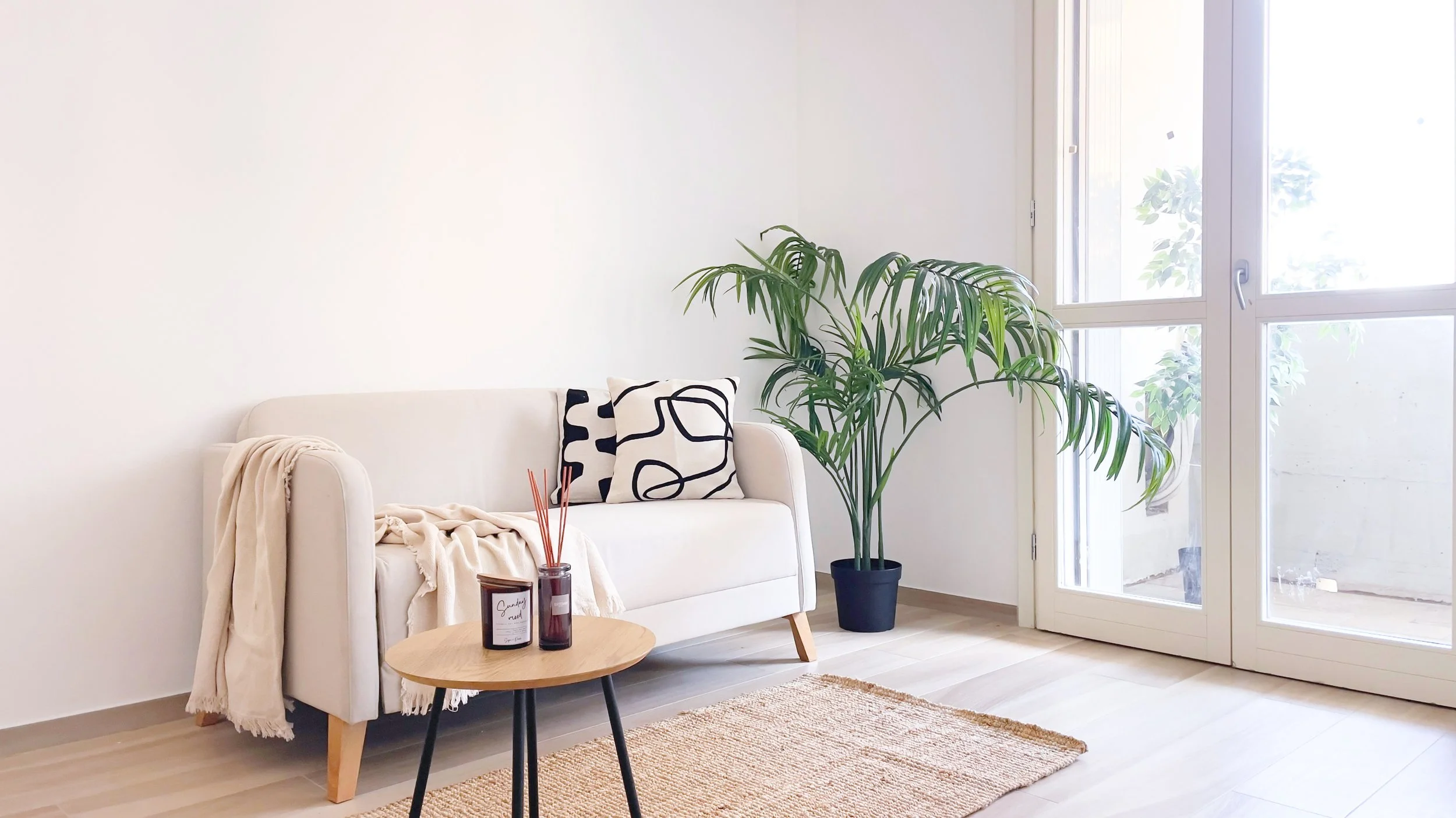Soggiorno luminoso con divano bianco, tavolino in legno, pianta in vaso, cuscini decorativi e grande finestra.