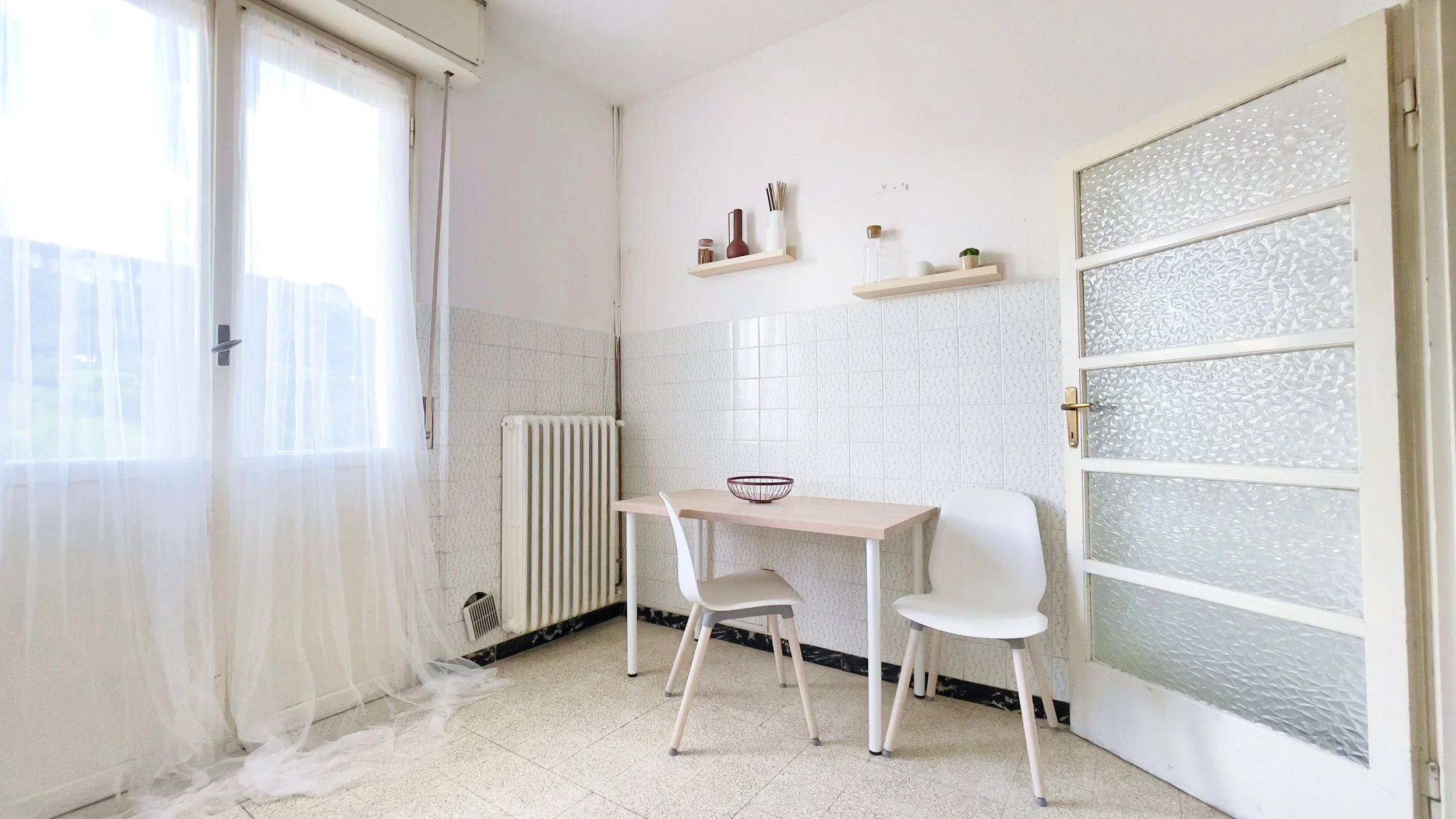 Stanza semplice e luminosa con tavolo e sedie moderne, mensole con decorazioni, porta vetro e finestra con tenda bianca.