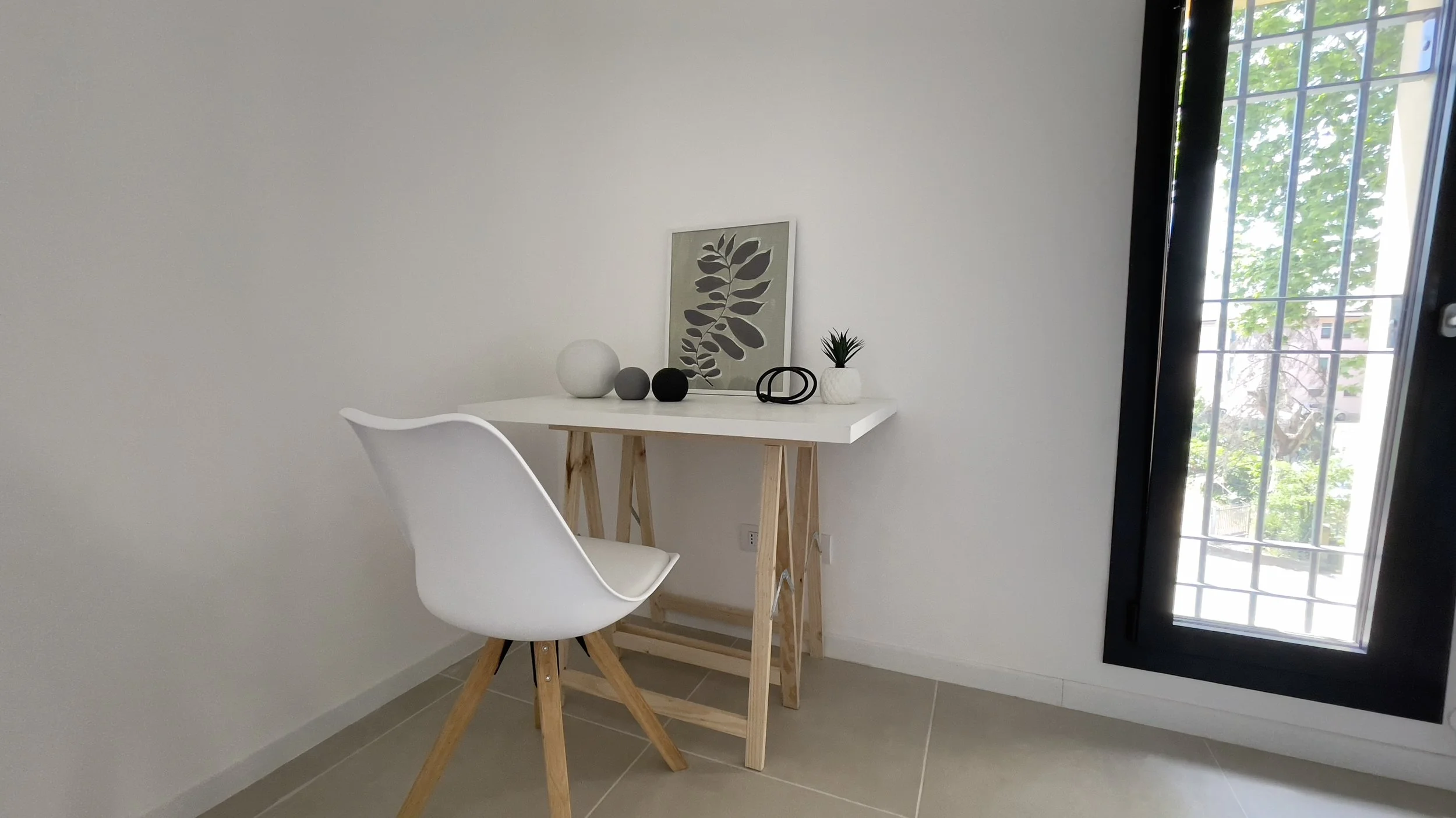 Scrivania minimalista con sedia bianca, decorazioni geometriche e pianta, vicino a finestra luminosa.