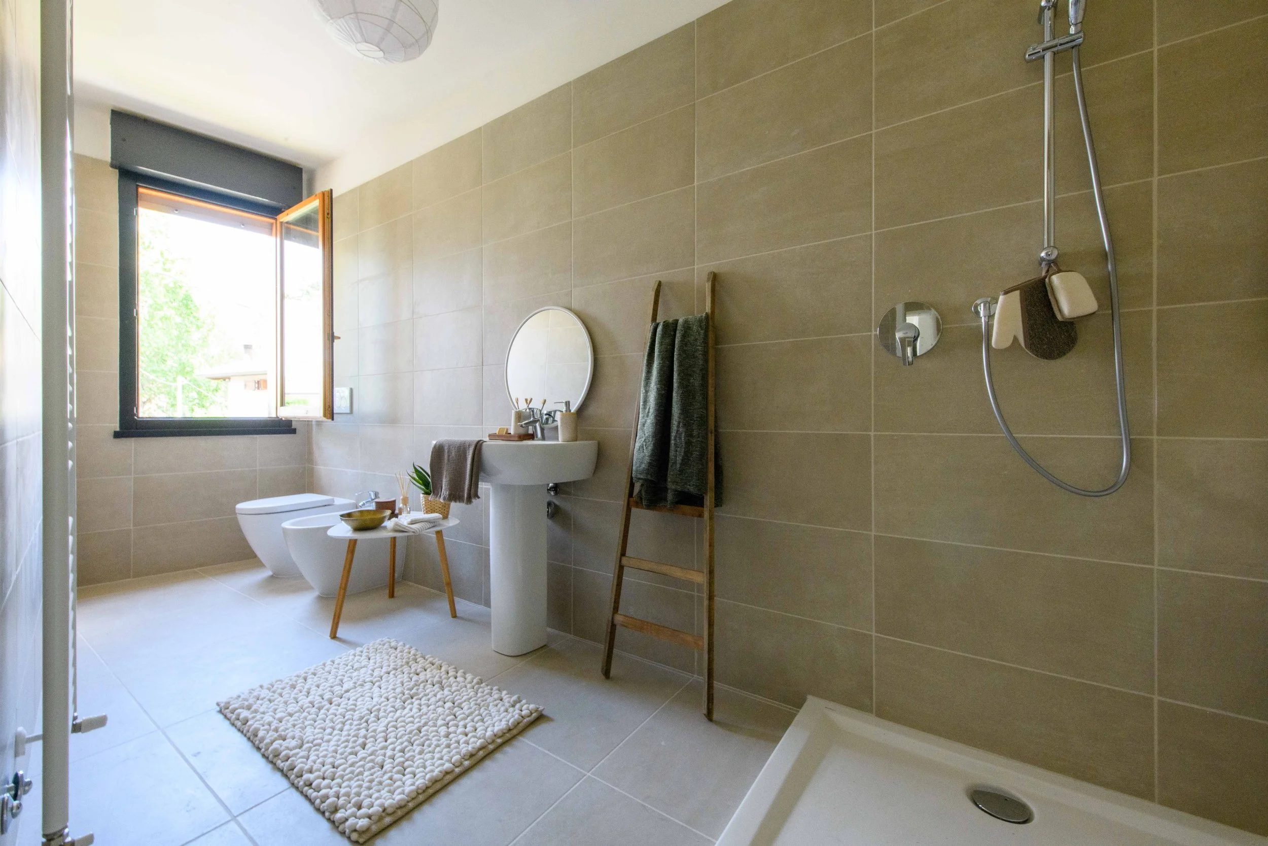 Bagno moderno con doccia, lavabo, bidet, e finestra aperta, decorato con tappetino e accessori in legno.