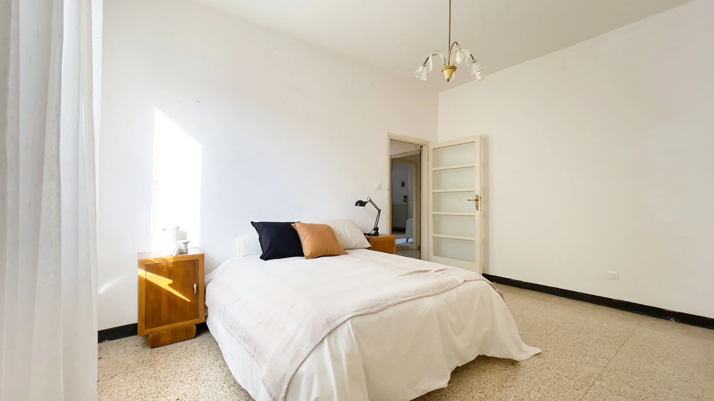 Camera da letto semplice e luminosa con letto matrimoniale, cuscini colorati, comodini in legno e lampada da tavolo, pavimento in piastrelle, pareti bianche e porta aperta.