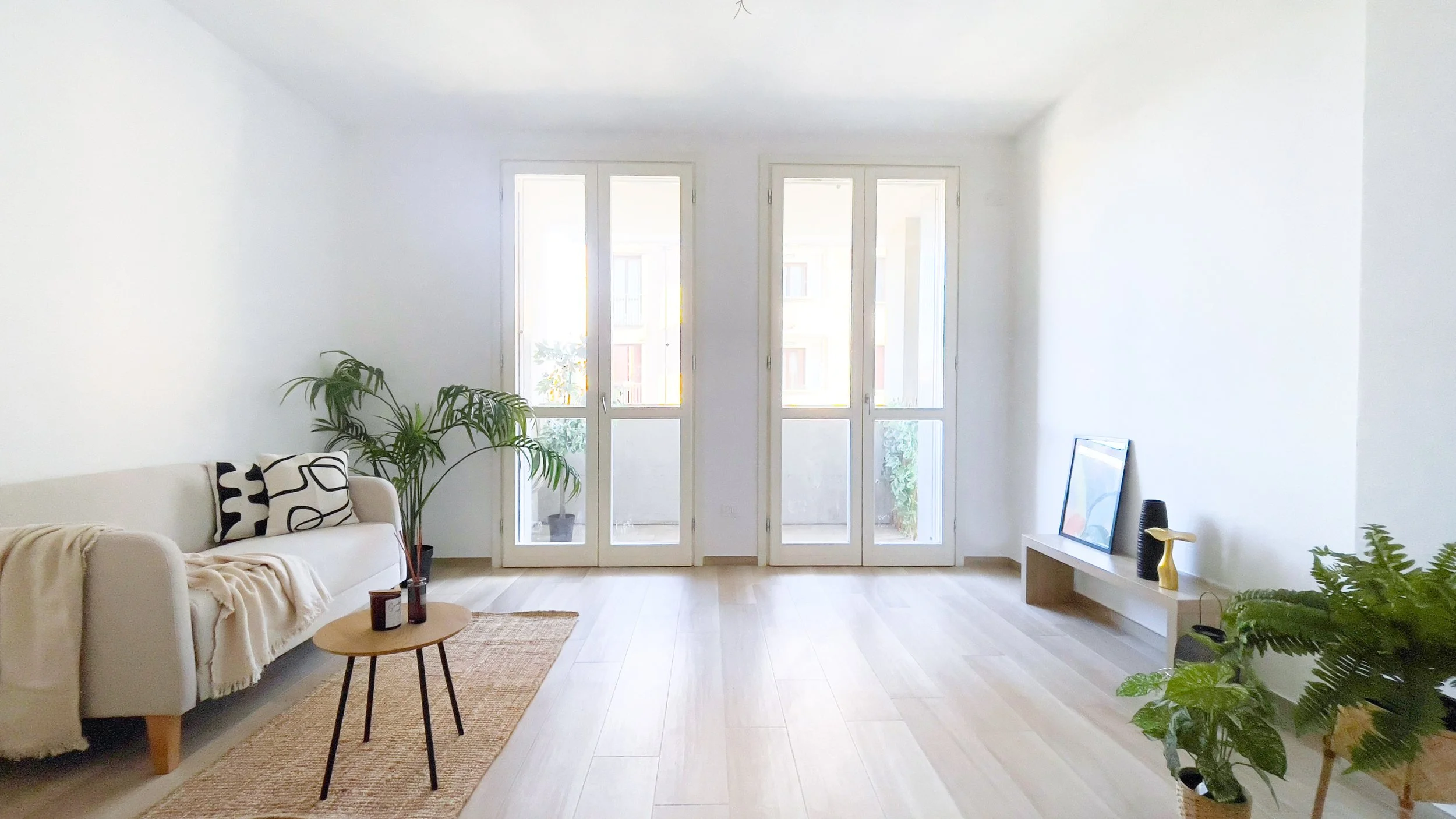 Soggiorno luminoso con divano beige, piante, tavolino e finestre grandi.