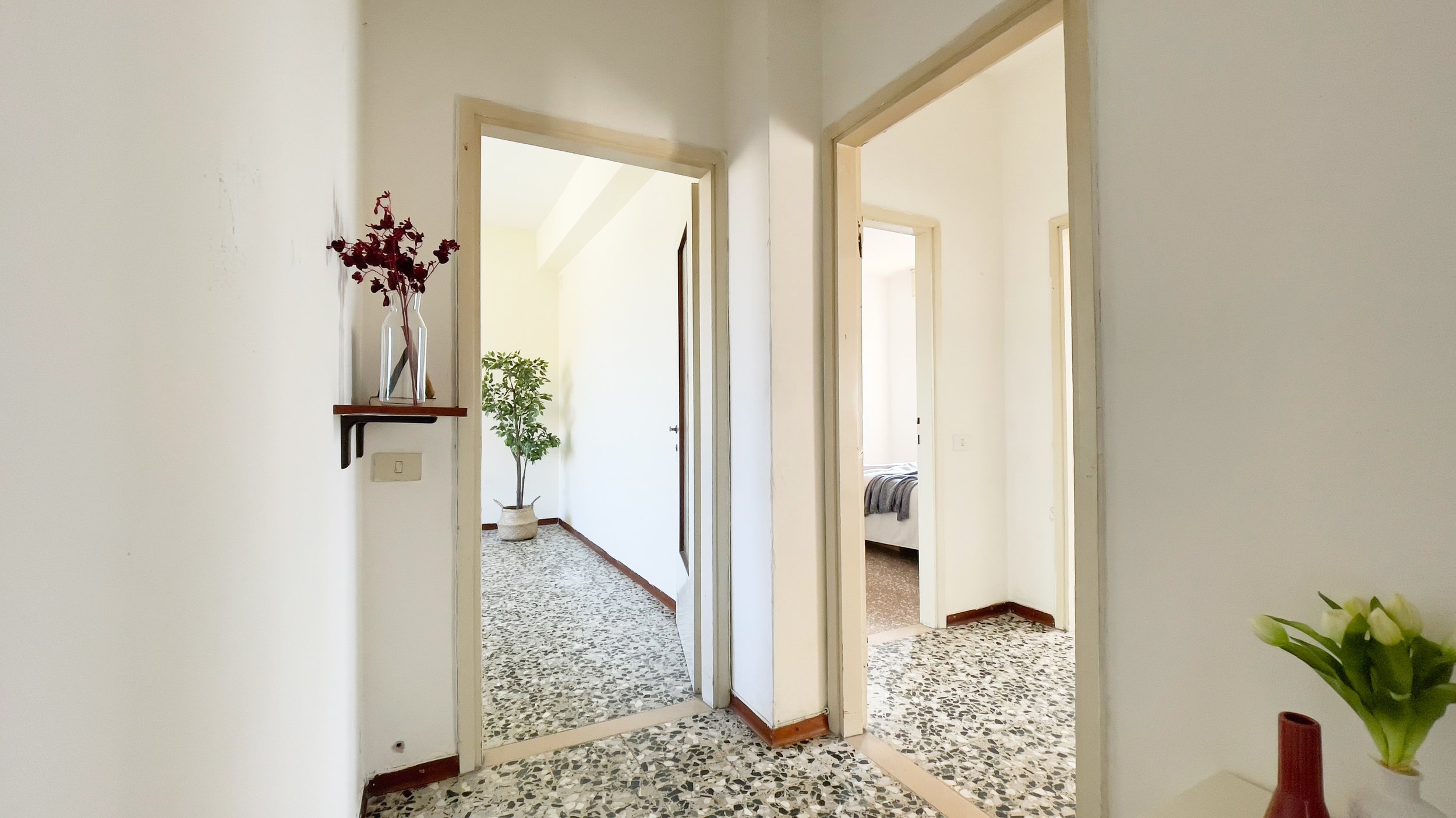 Corridoio di un appartamento con pavimento in marmo, due porte aperte, una mensola con fiori rossi e un vaso con fiori bianchi su un mobile.