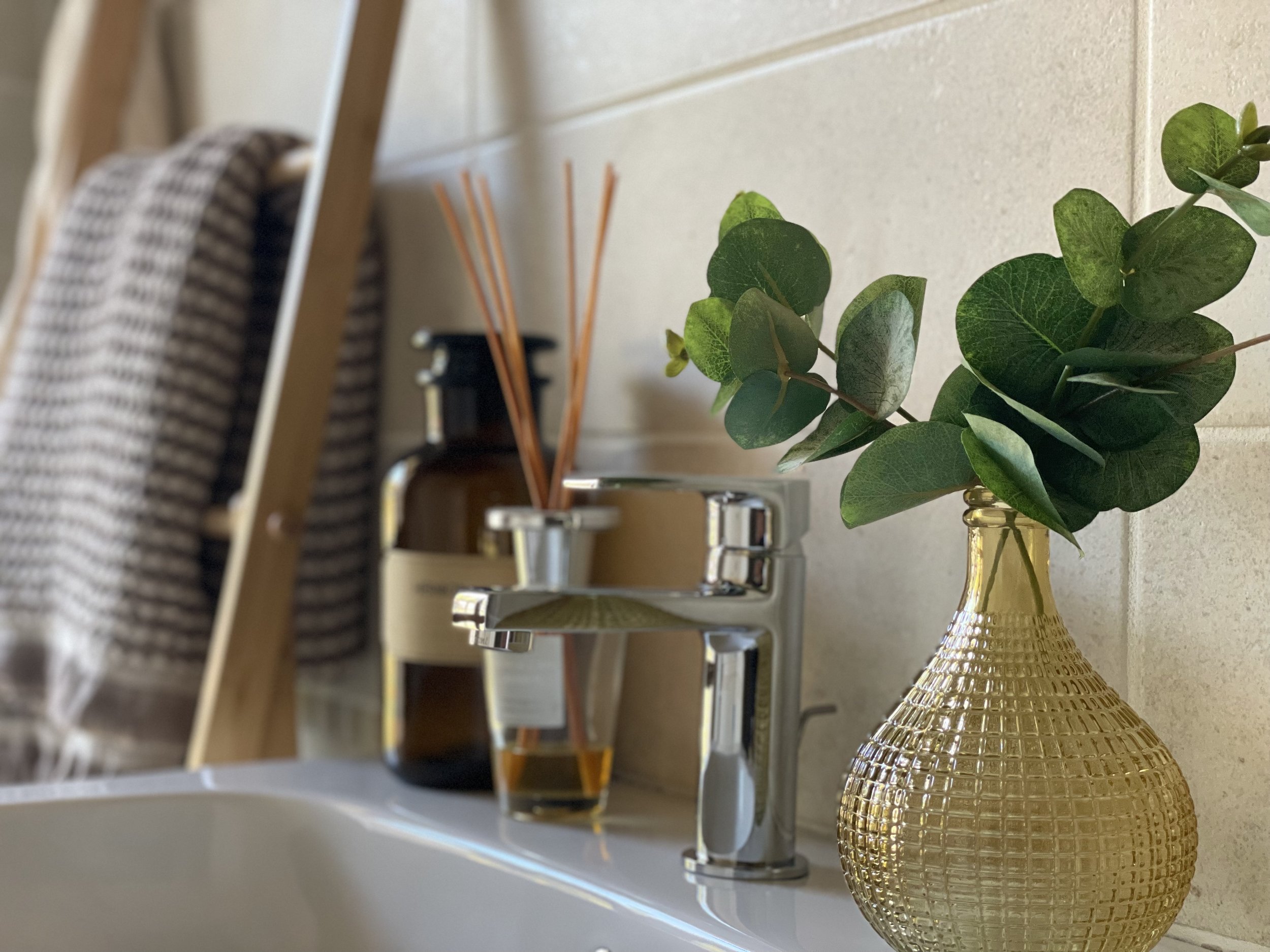 Bagno con rubinetto cromato, vaso di vetro con foglie verdi, diffusore di profumo e asciugamani su scala in legno.