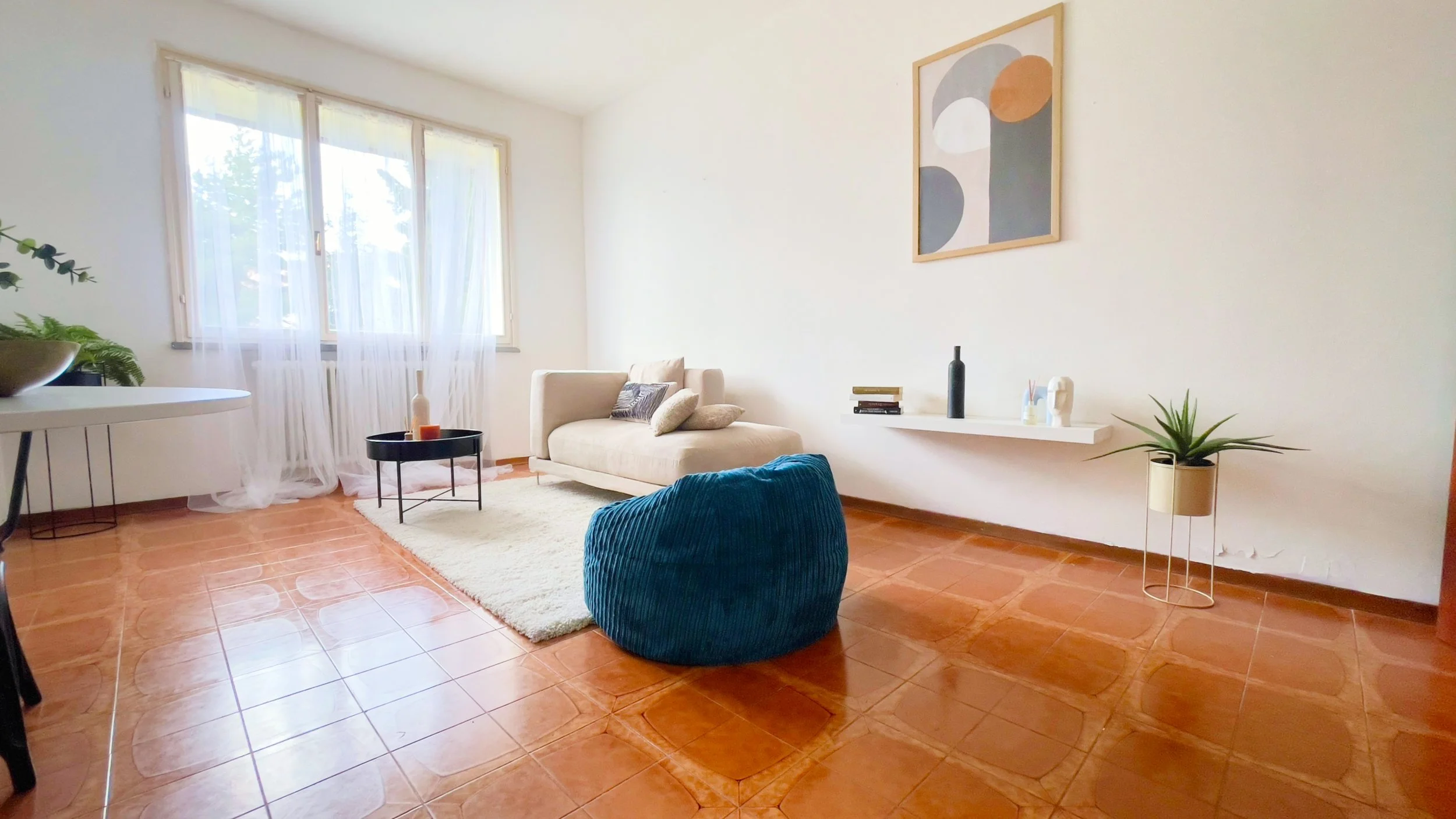 Soggiorno moderno con divano beige, poltrona blu, tavolino rotondo nero, tappeto bianco, pianta in vaso, tavolo bianco, e finestra con tende trasparenti.