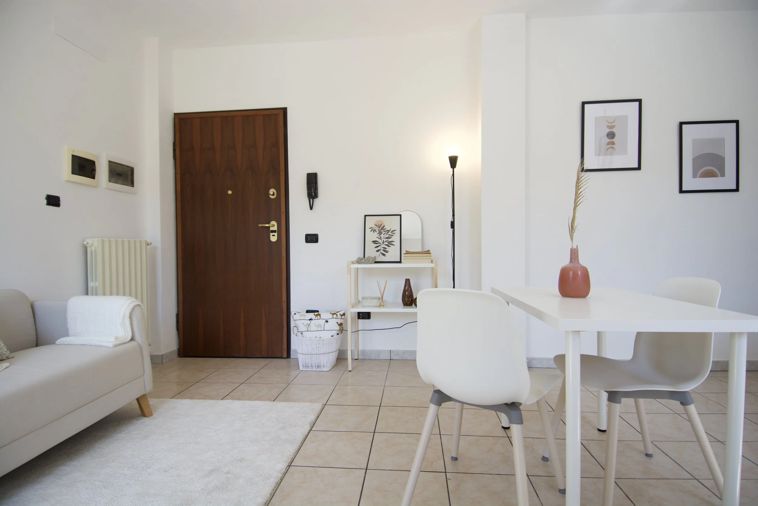Interno di un salotto moderno e minimalista con porta d'ingresso in legno, un divano chiaro con coperta, tavolo bianco con sedie, decorazioni a parete, scaffale con cornice e lampada.