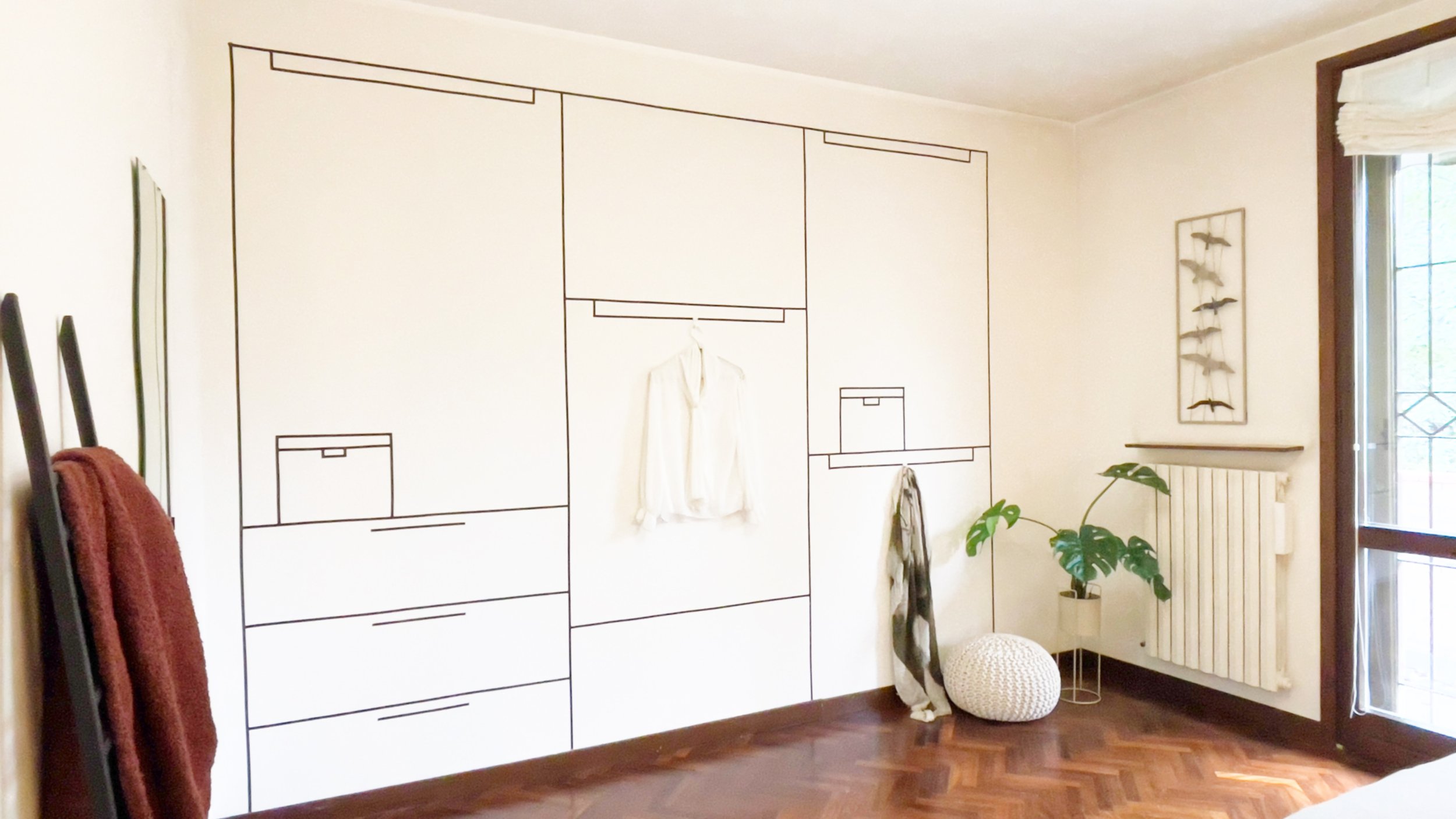 Una stanza con un armadio stilizzato dipinto sul muro. Una camicia bianca è appesa al centro del disegno. Accanto ci sono un radiatore, una pianta verde e un pouf intrecciato bianco. Sul lato sinistro ci sono due asciugamani appesi su un supporto.